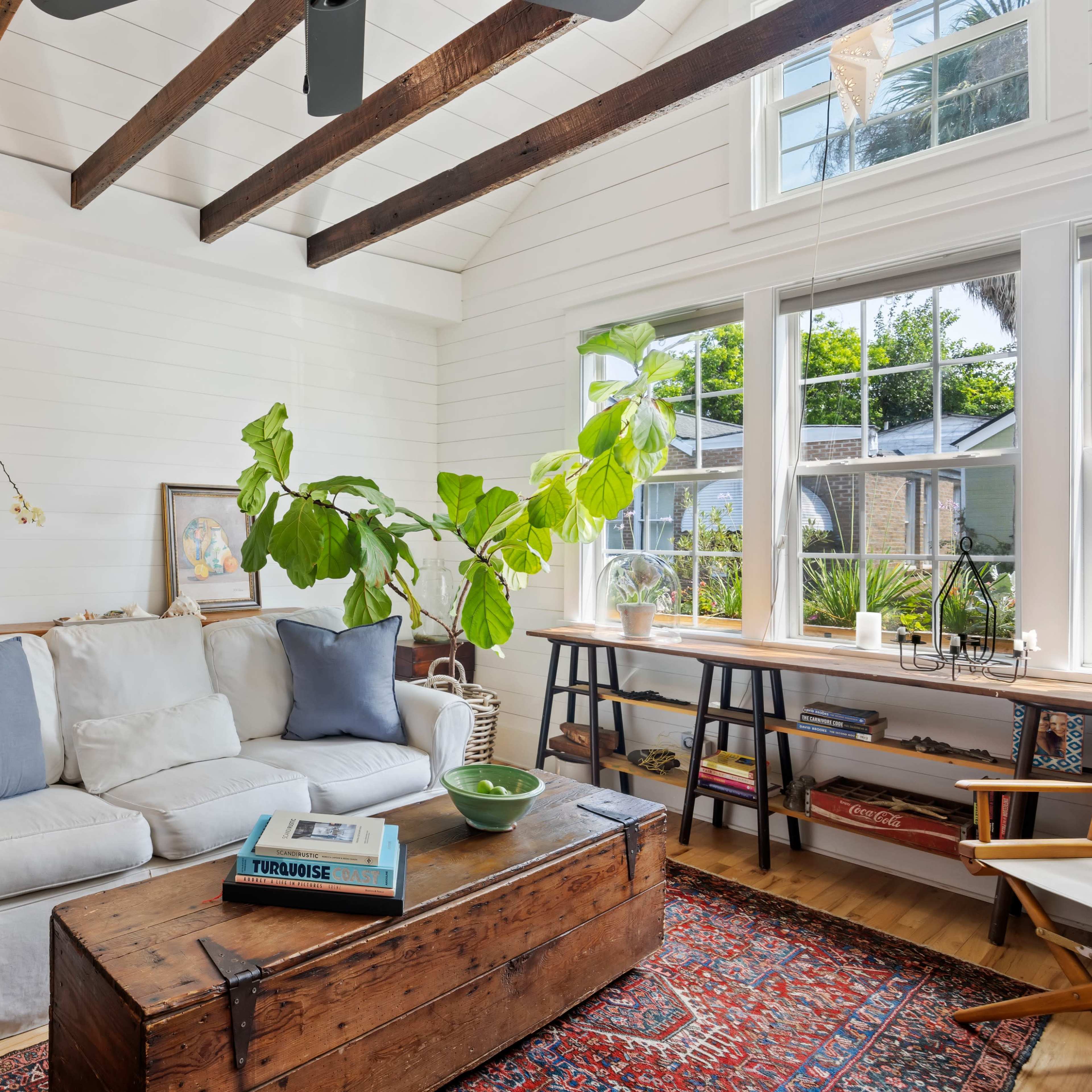 A bright living room features a white sofa with blue cushions, a wooden coffee table, and a large plant by the window.