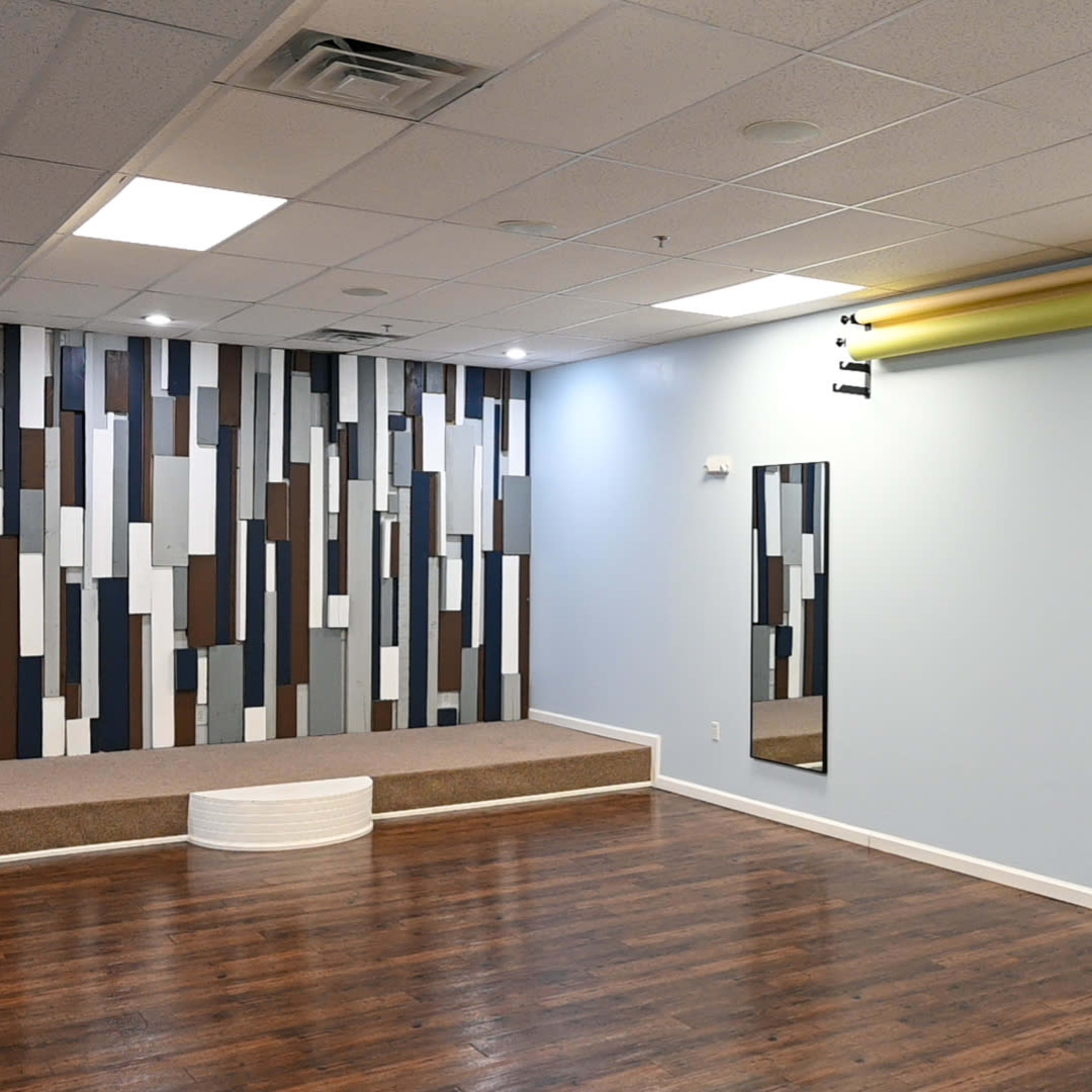 A spacious room with a hardwood floor, featuring a decorative wall of vertical stripes, two mirrors, and a small raised platform.