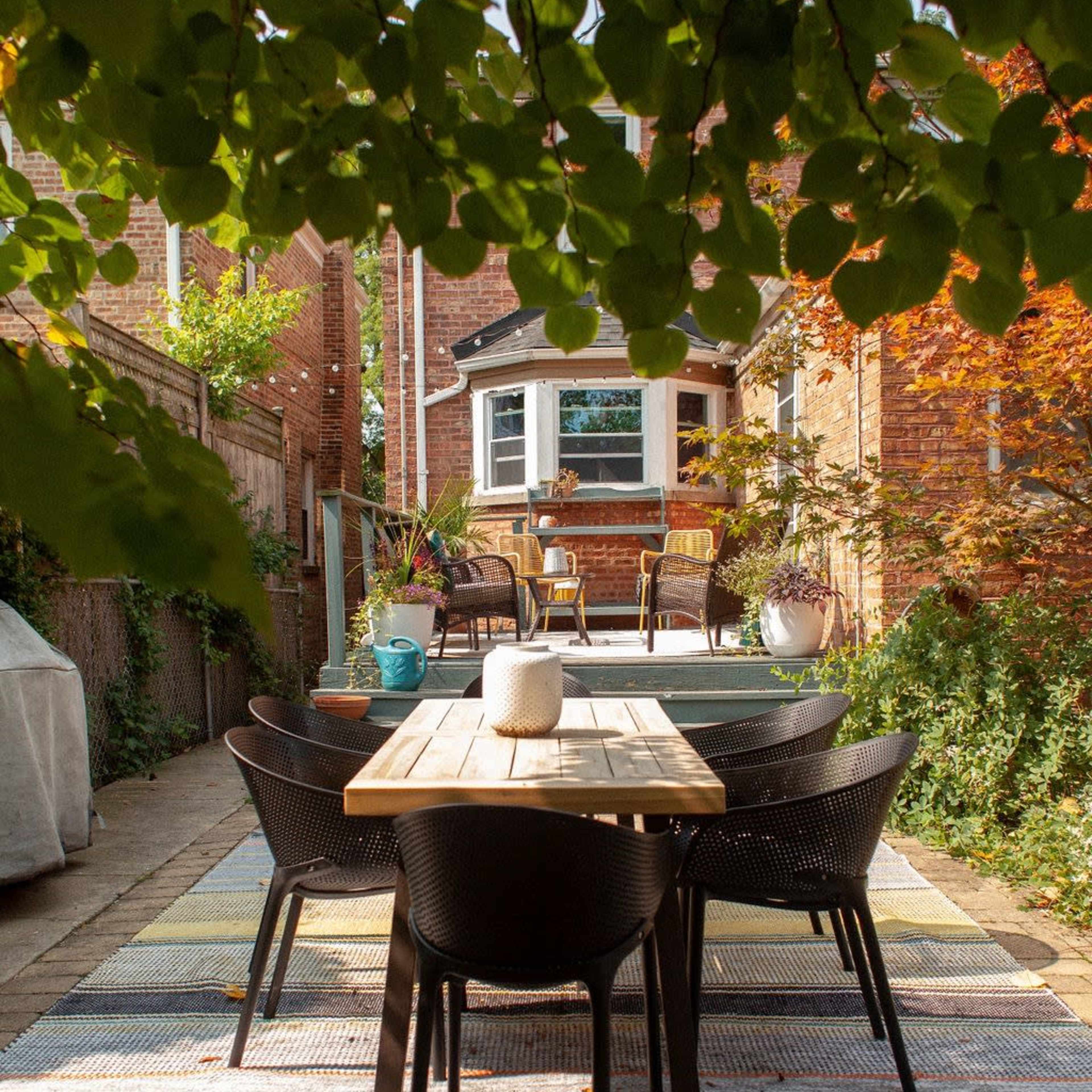 A wooden dining table surrounded by black chairs is set on a patterned rug in a backyard, with plants and a seating area visible in the background.
