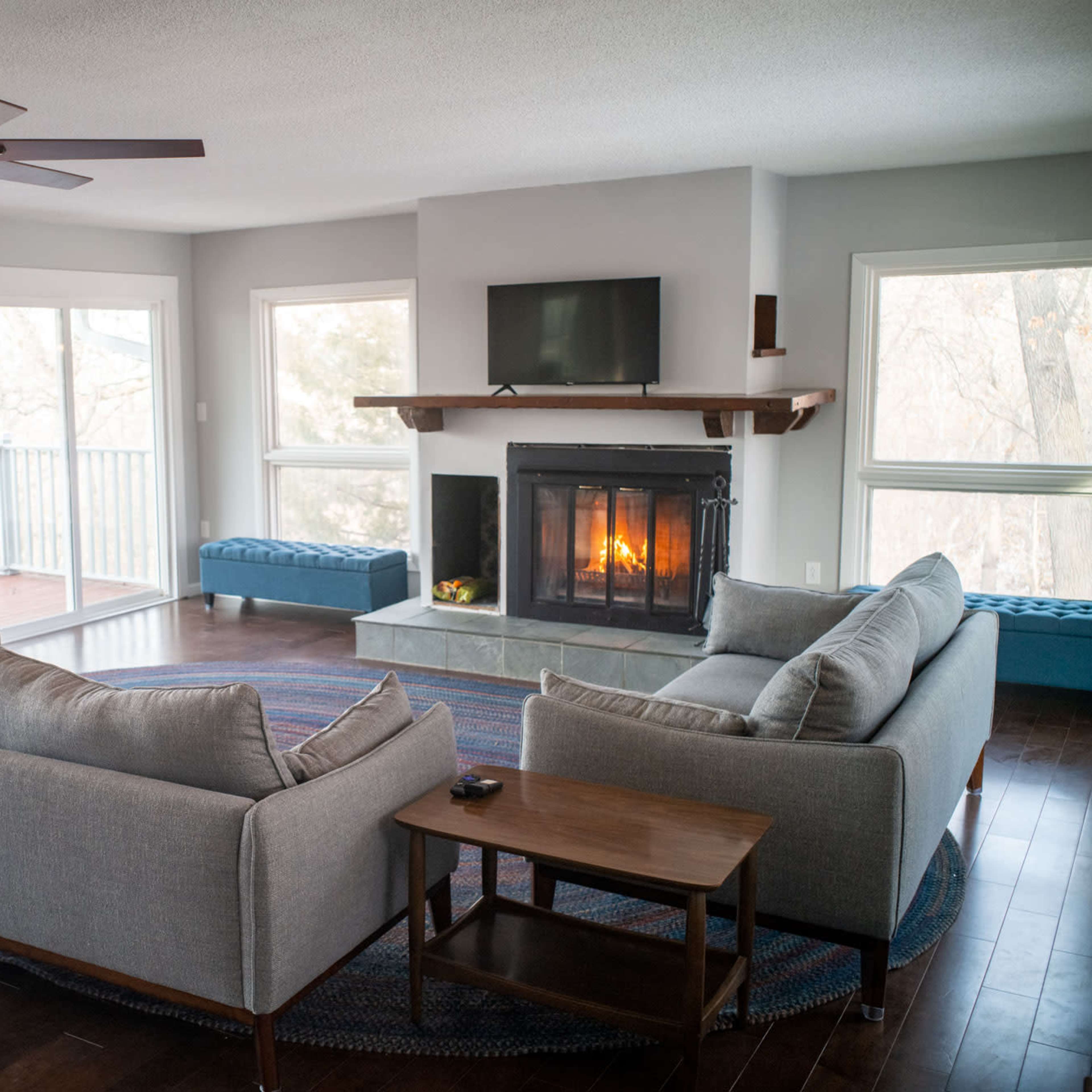A modern living room features a sofa set, a coffee table, and a fireplace with a television above it, all surrounded by large windows.
