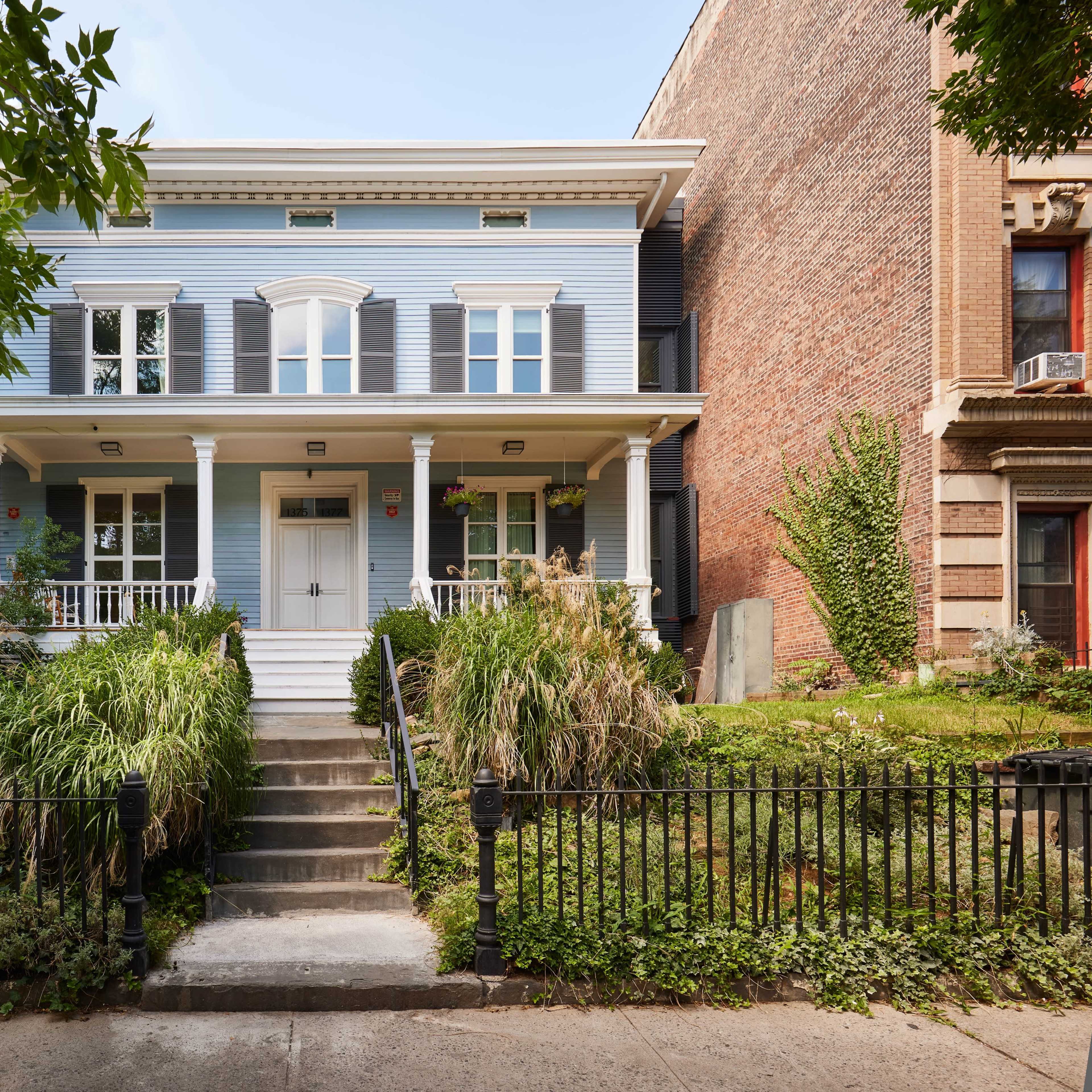 A two-story blue house with white columns and a front porch, surrounded by a landscaped yard and a wrought-iron fence.