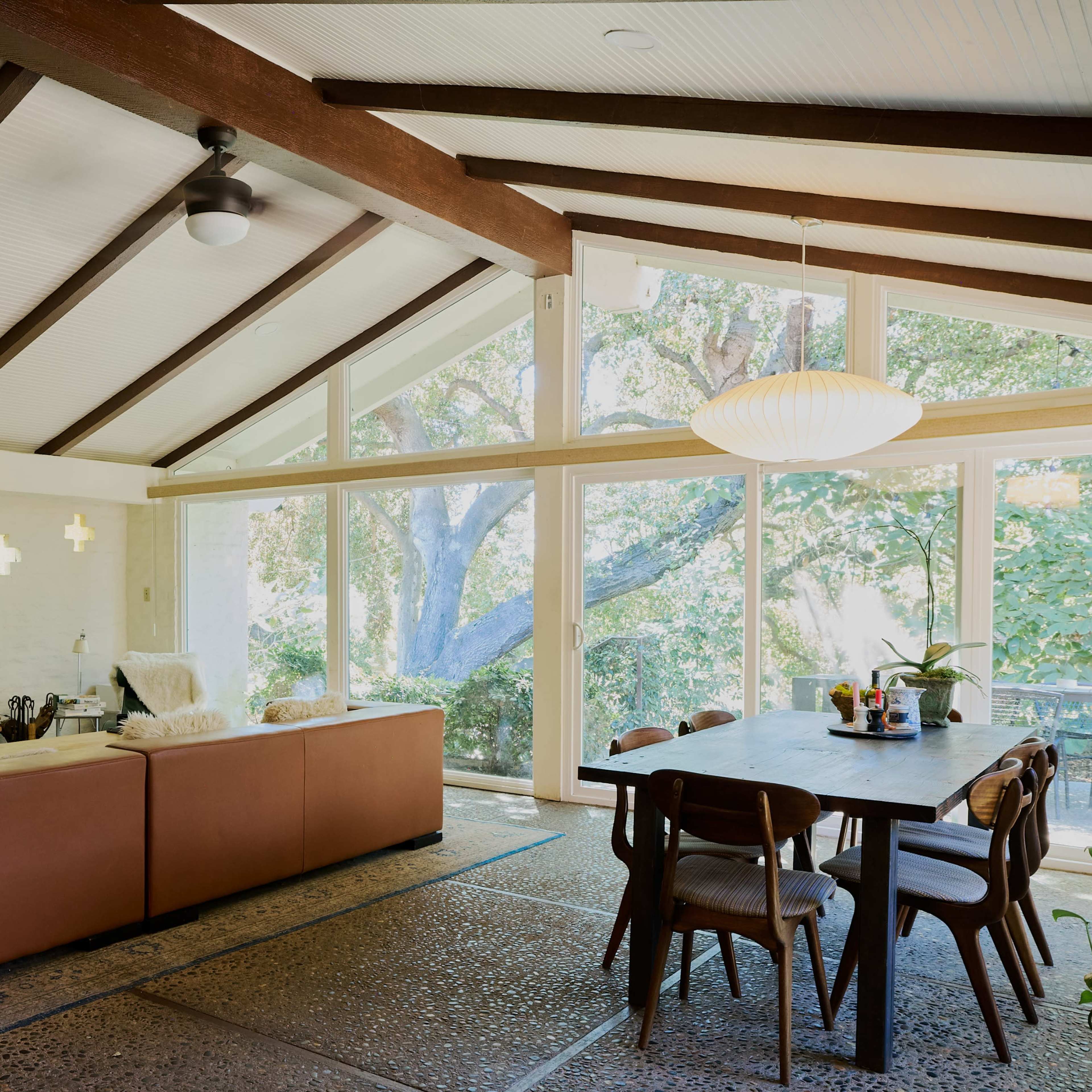 A spacious living area with a brown sectional sofa, a wooden dining table, and large windows overlooking a tree-filled outdoor space.