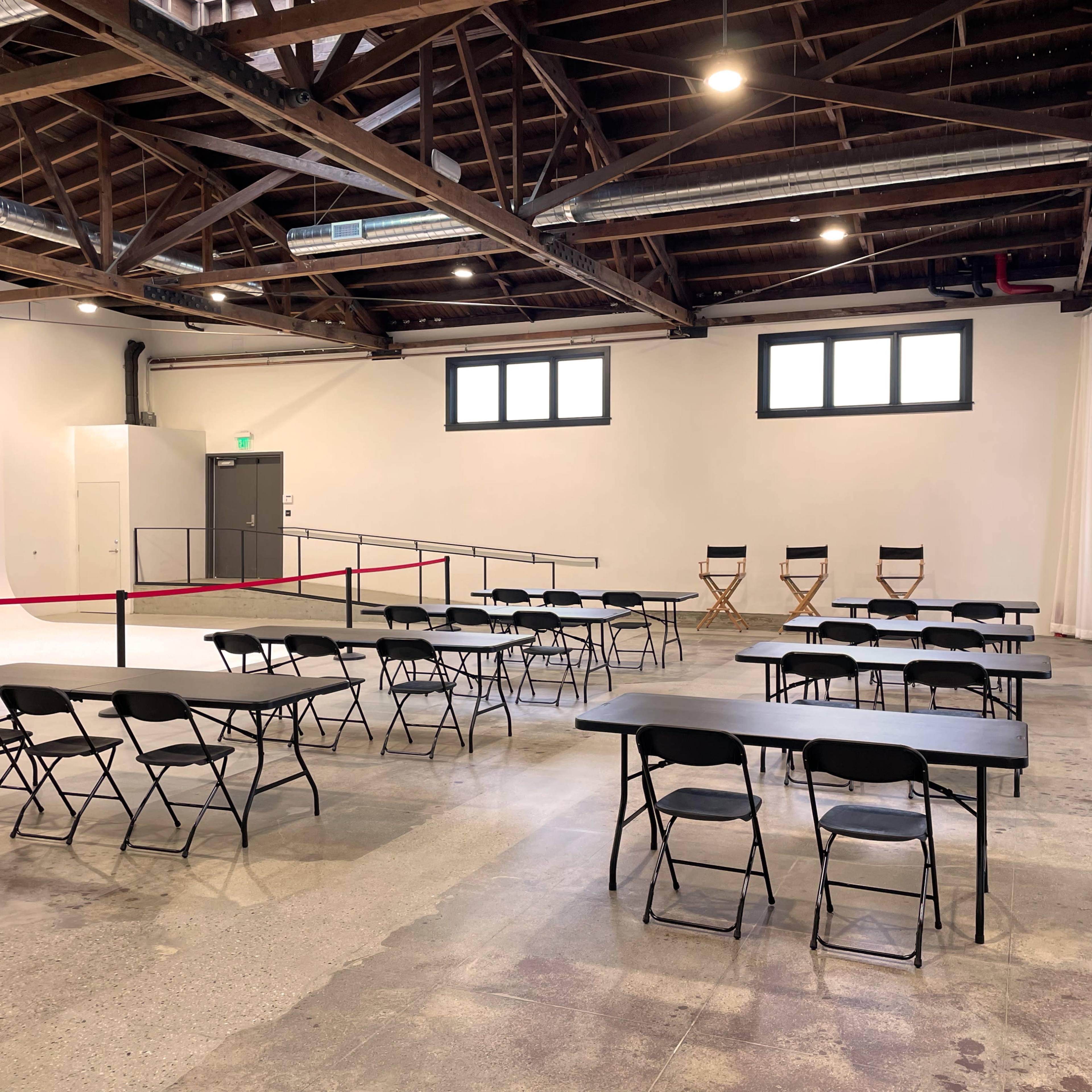 A spacious room with several rows of black tables and chairs, set up for an event, under a wooden beam ceiling.