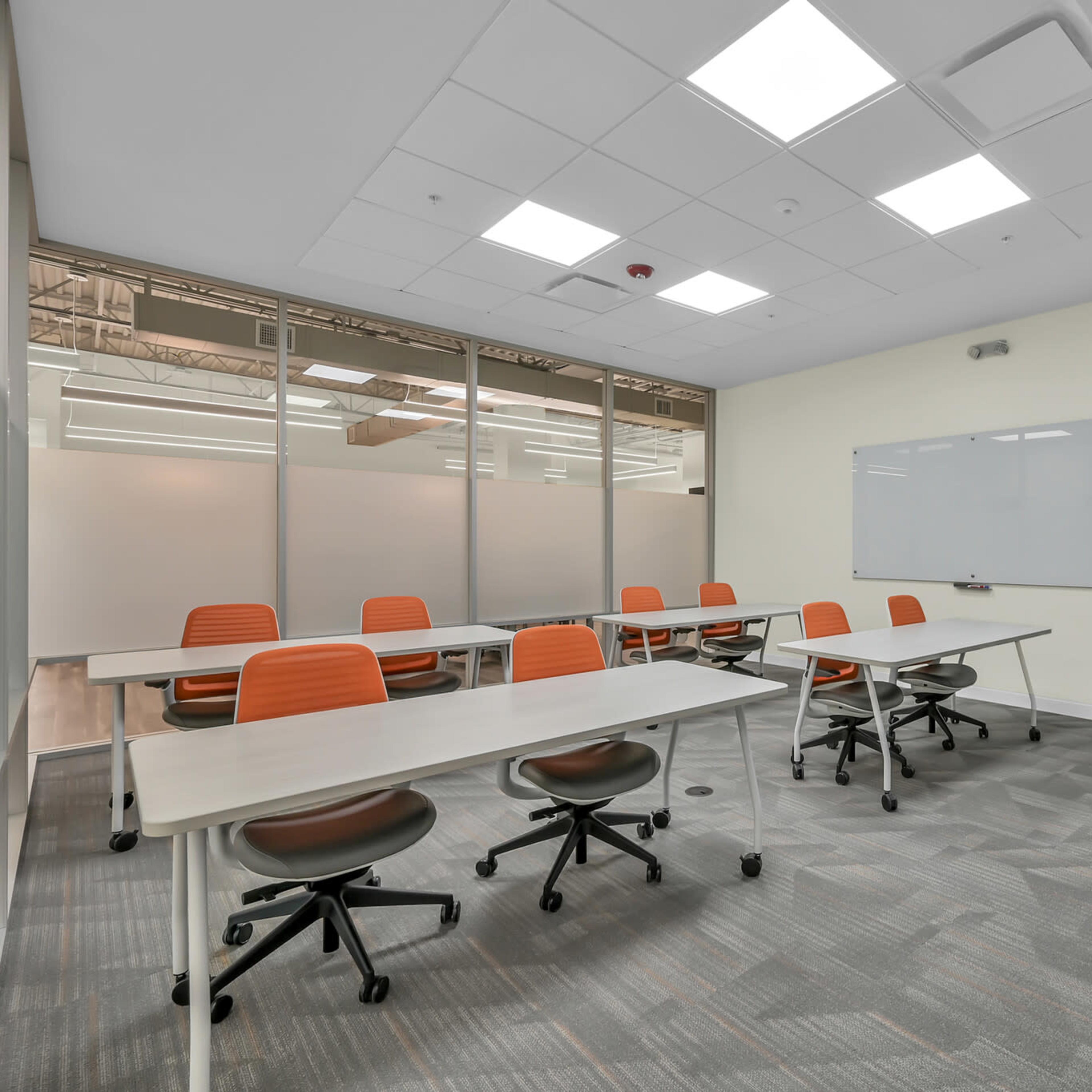 A modern classroom features several tables with orange chairs, a whiteboard, and large windows along one wall.
