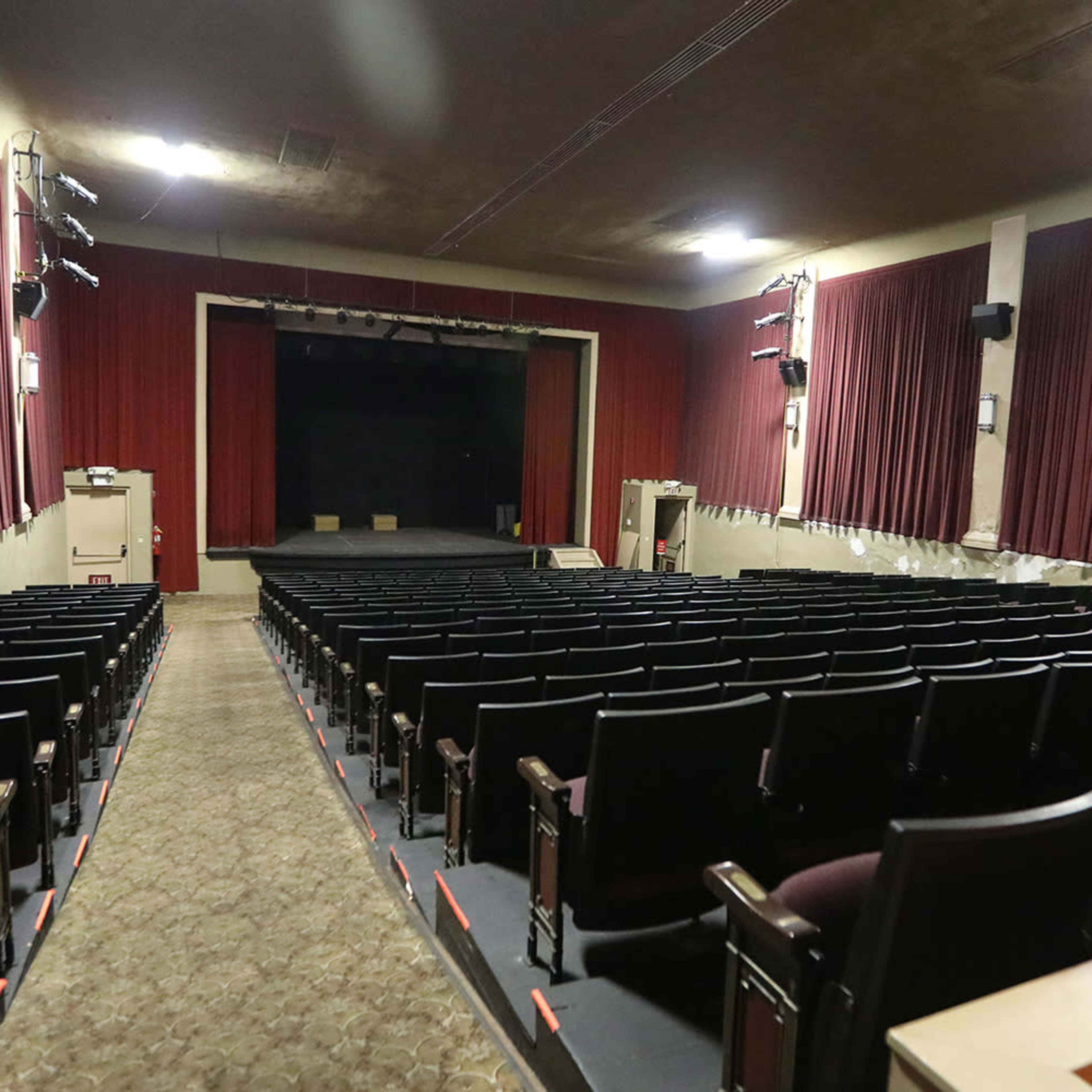 An empty theater with rows of black seats facing a stage, framed by red curtains and dim lighting.
