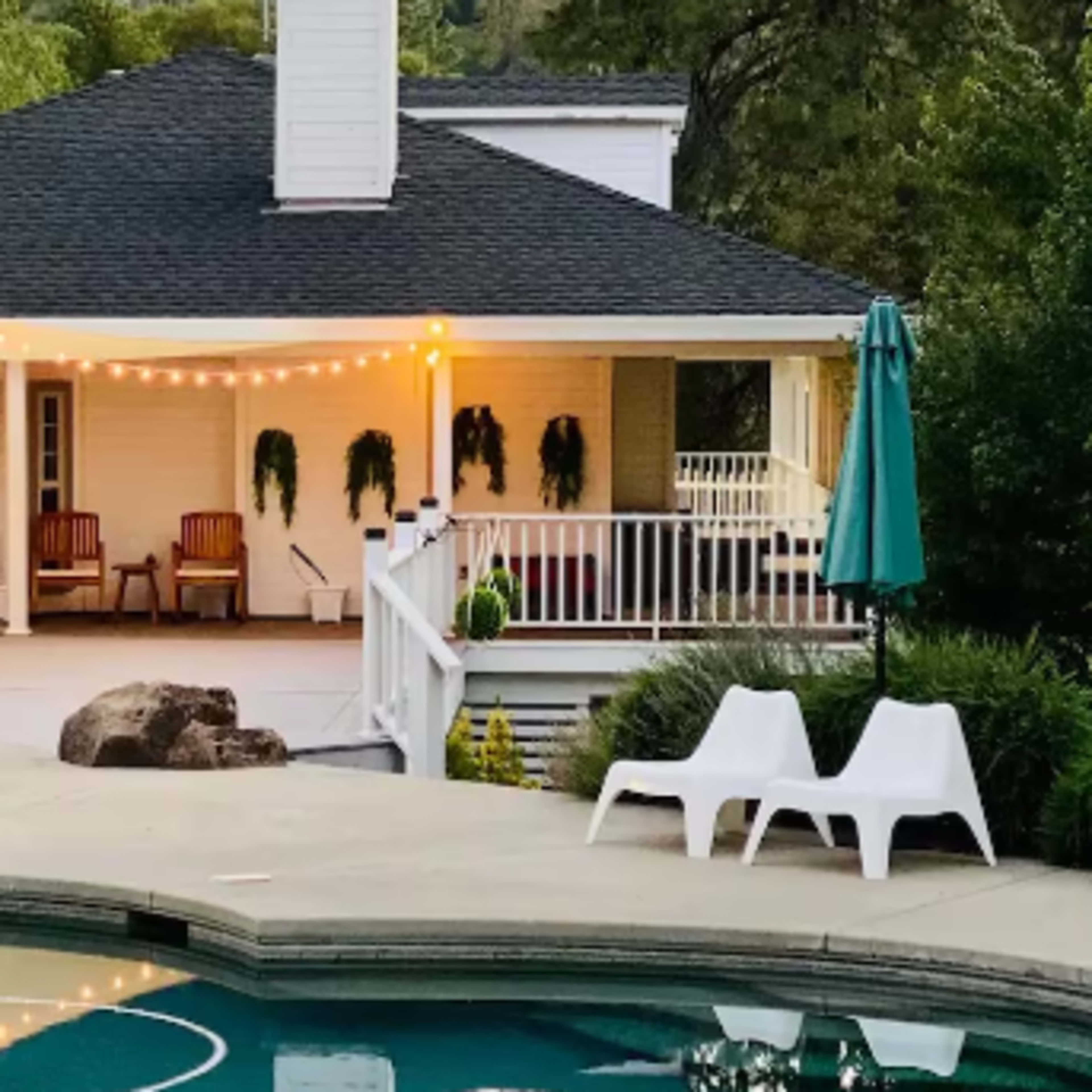 A backyard with a swimming pool, patio area with seating, and a house featuring a porch with string lights and plants.