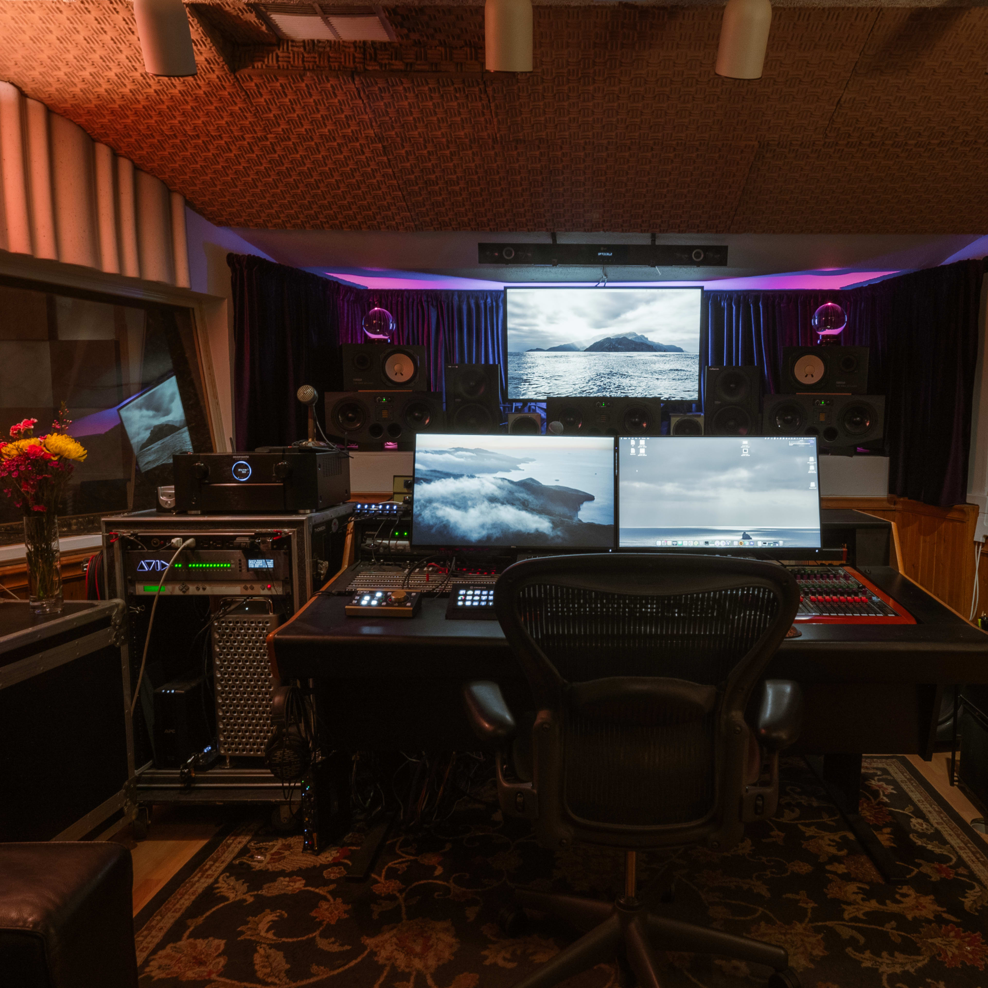 A music studio with a central workstation featuring two large screens, surrounded by audio equipment, speakers, and a guitar resting on a chair.