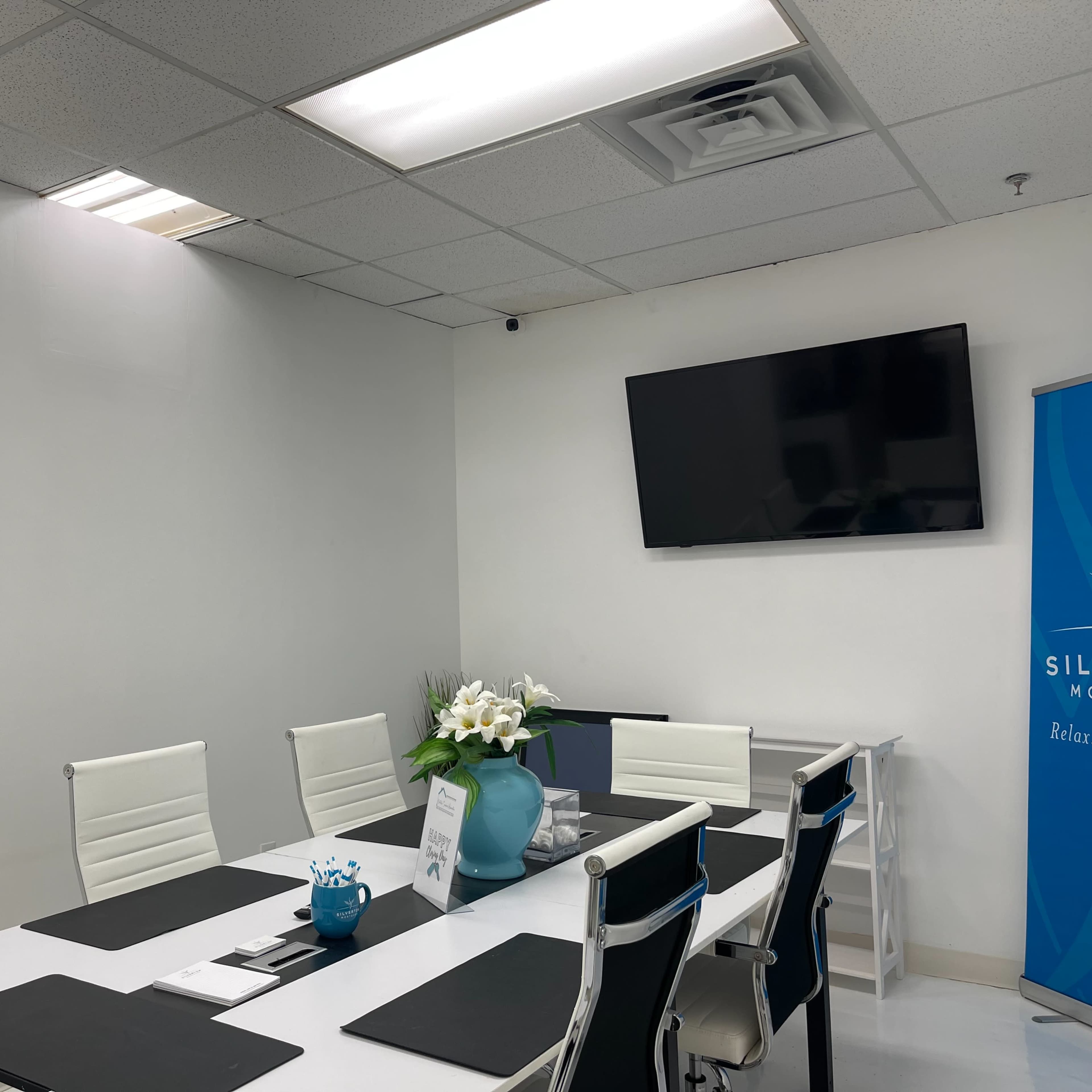 A modern conference room features a rectangular table surrounded by white chairs, with a television mounted on the wall and a vase of flowers in the center.