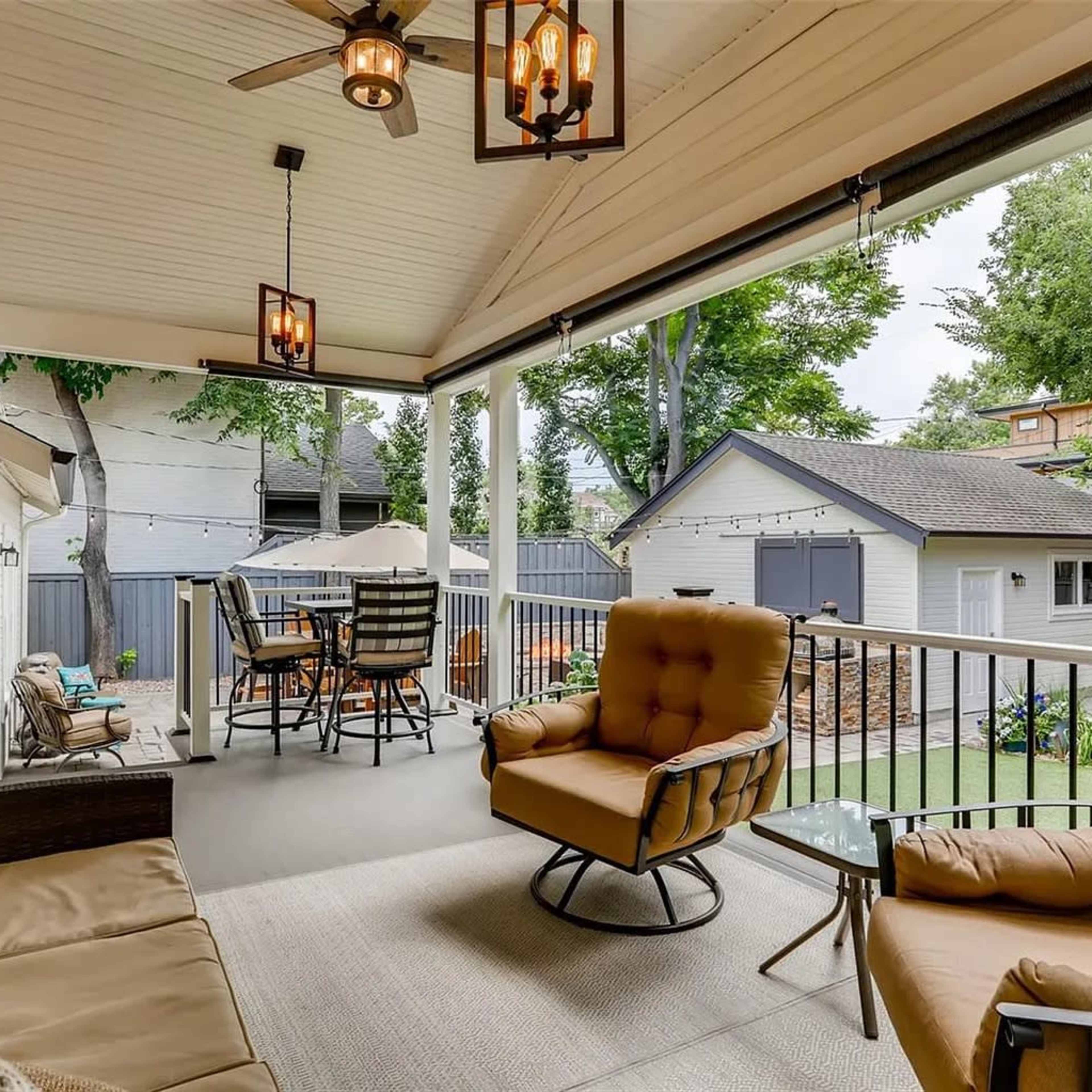 A covered porch area with a seating arrangement, a TV mounted on the wall, and a view of a backyard with a garage in the background.