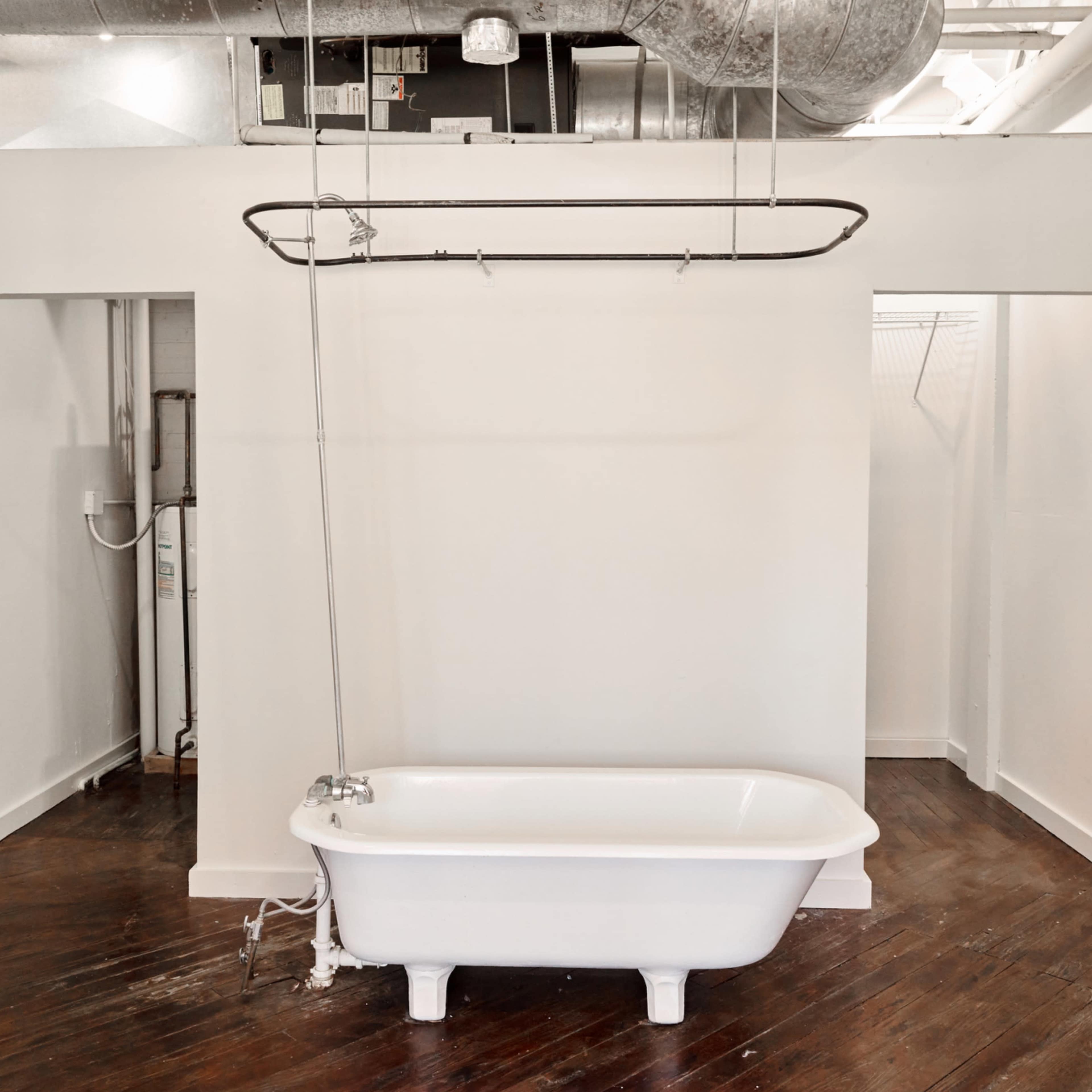 A minimalist bathroom space featuring a white bathtub with a metal shower rod suspended above it, surrounded by plain white walls and dark wooden flooring.