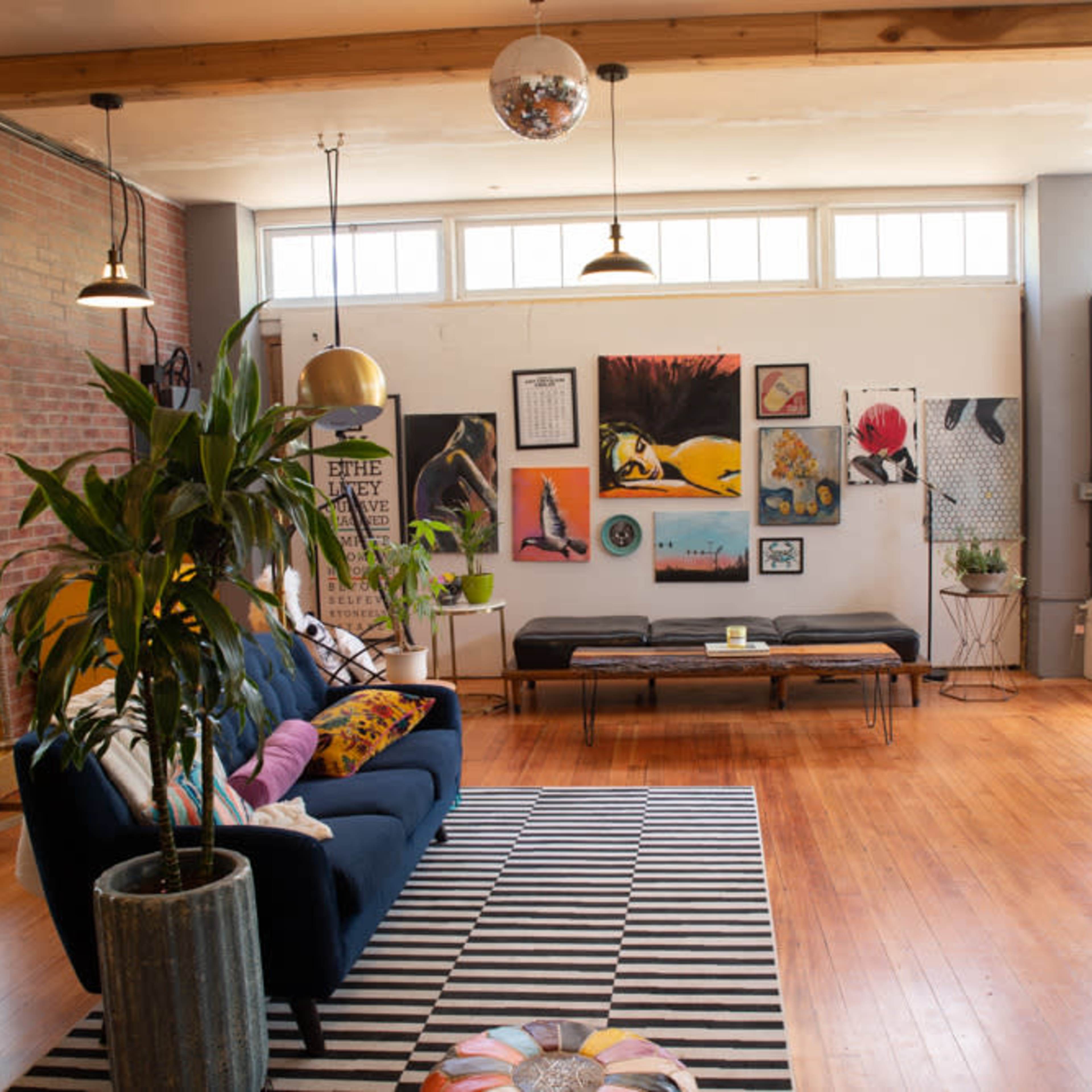 A bright living area featuring a blue sofa, a striped rug, and various art pieces on the walls, illuminated by natural light from large windows.