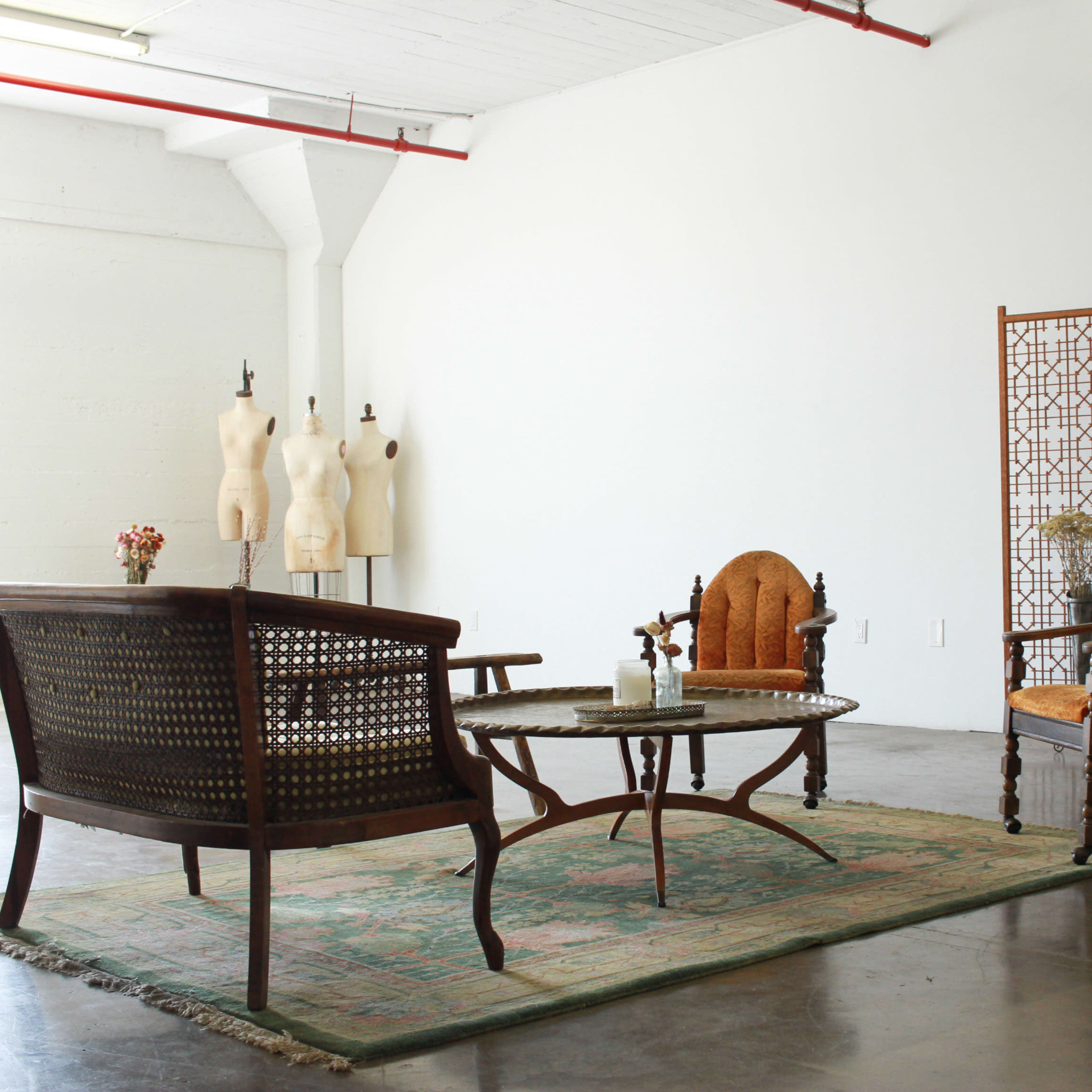 A minimalist room with a wooden seating arrangement, a round table, and decorative screens, complemented by a vintage area rug and dressmaker forms in the background.