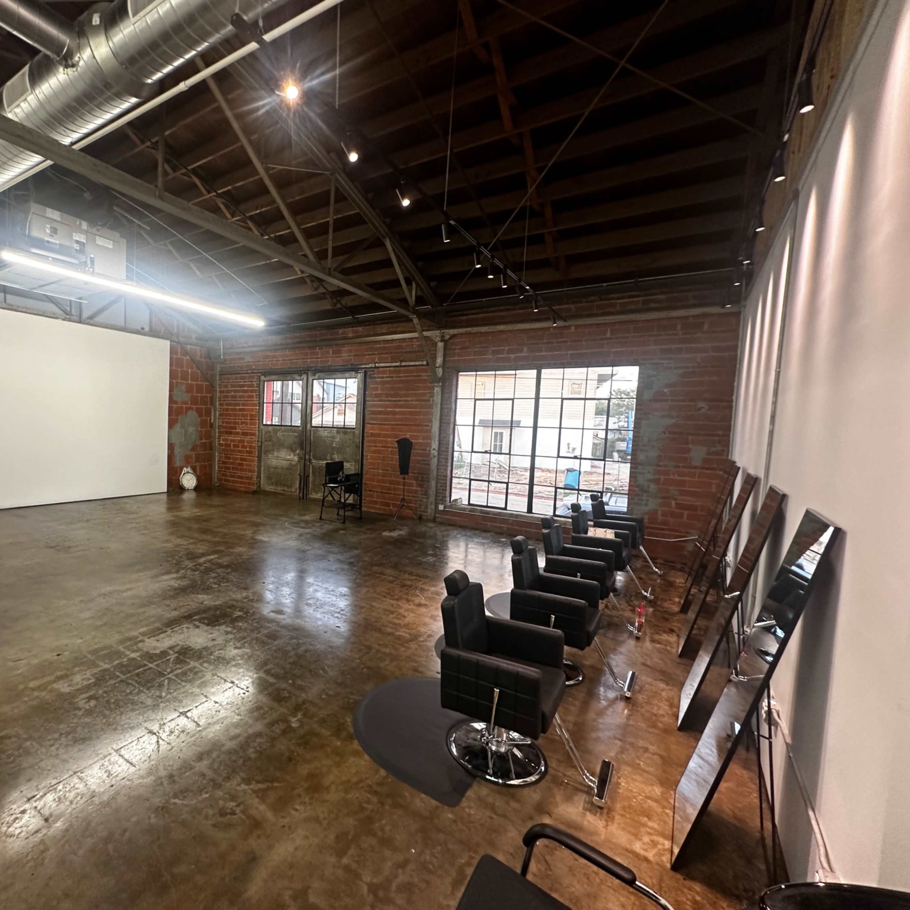 An open, spacious studio with a polished concrete floor, exposed brick walls, and several black salon chairs lined up against the wall.