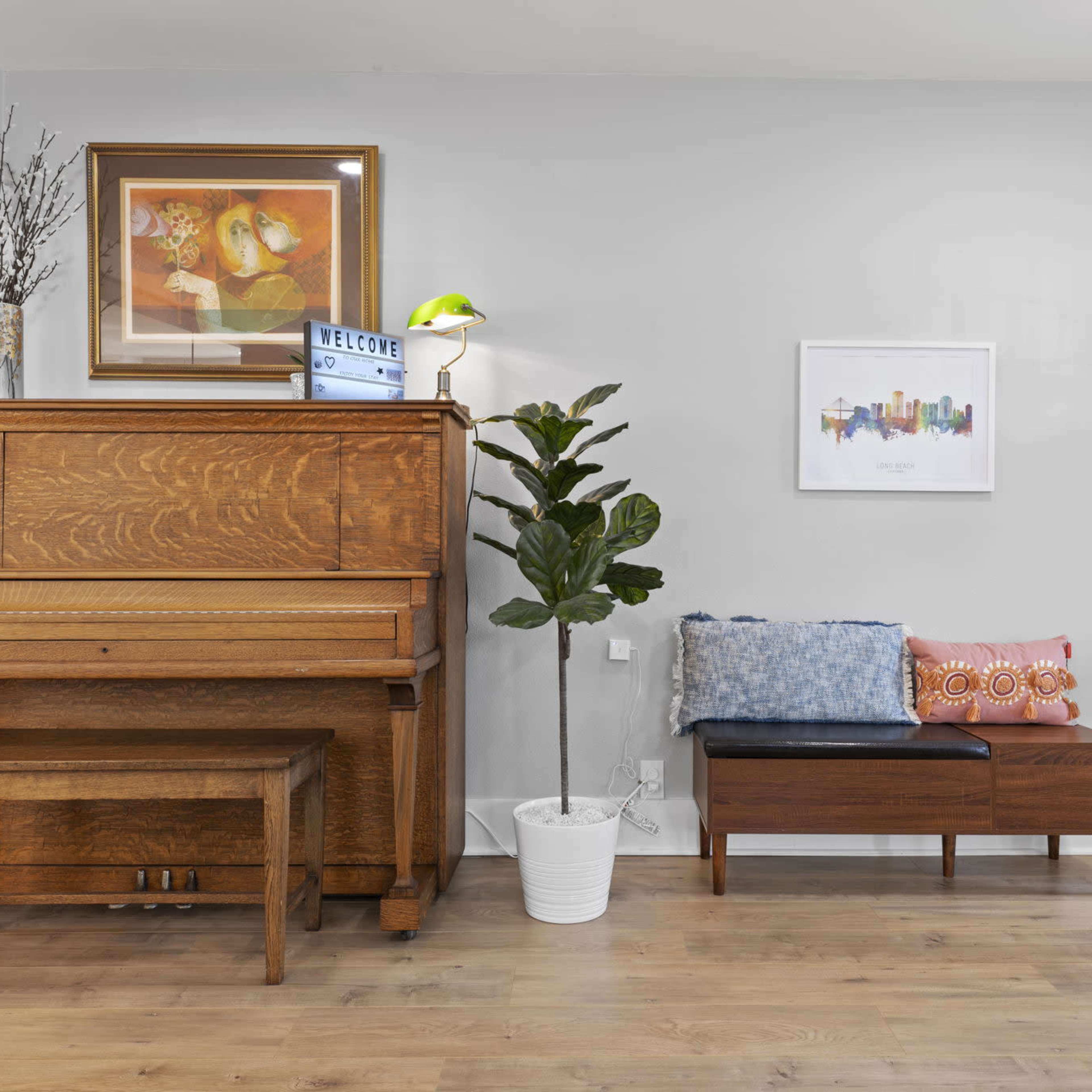 A wooden piano sits against a light gray wall beside a potted plant and a coat rack, while a bench decorated with pillows is positioned nearby.