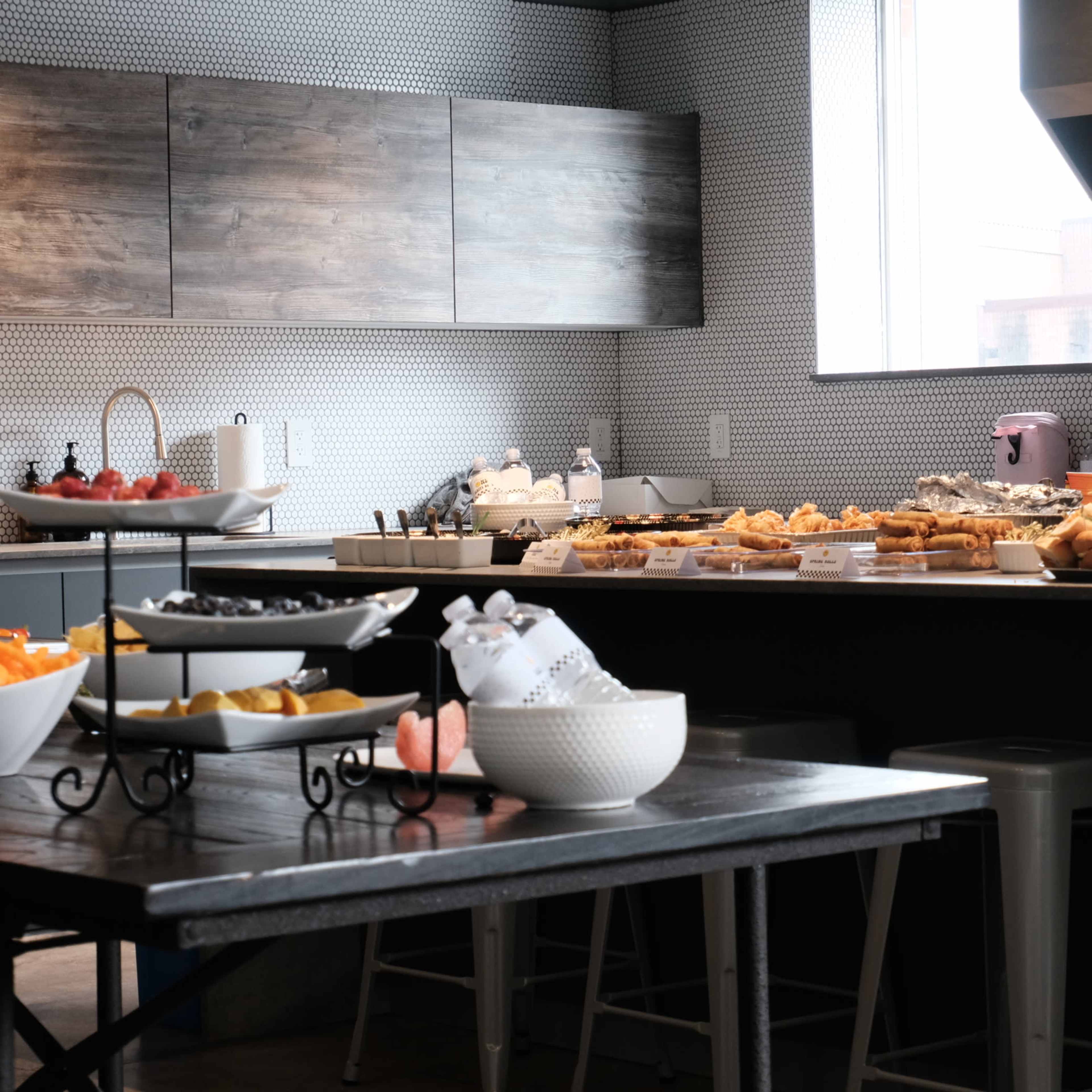 A buffet table filled with various food items is set up in a modern kitchen space with light-colored walls and a large window.