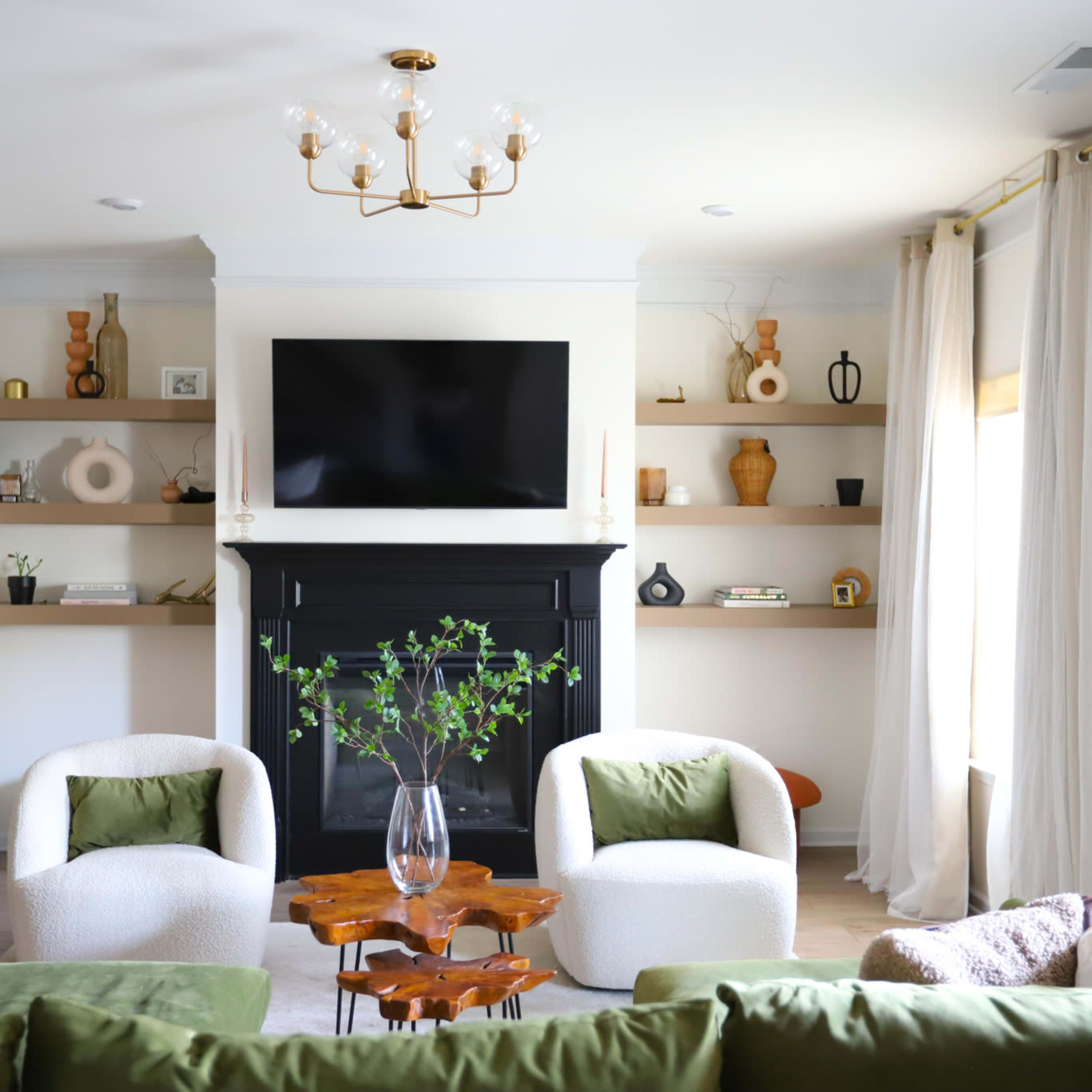 A modern living room features two upholstered chairs, a sofa, a black fireplace, and shelves with decorative items and plants.