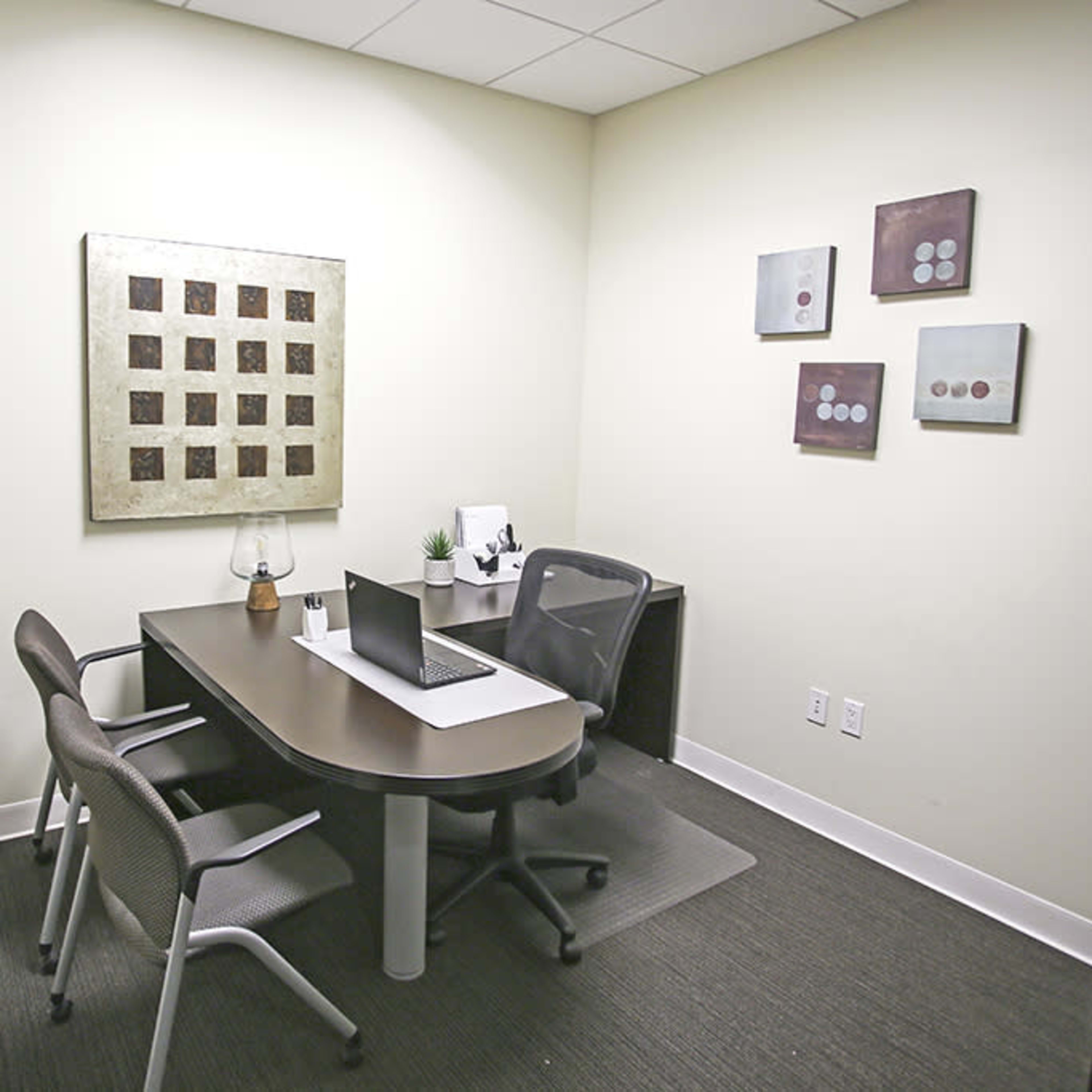 A small office space featuring a desk with a laptop, two chairs, a plant, and framed wall art.