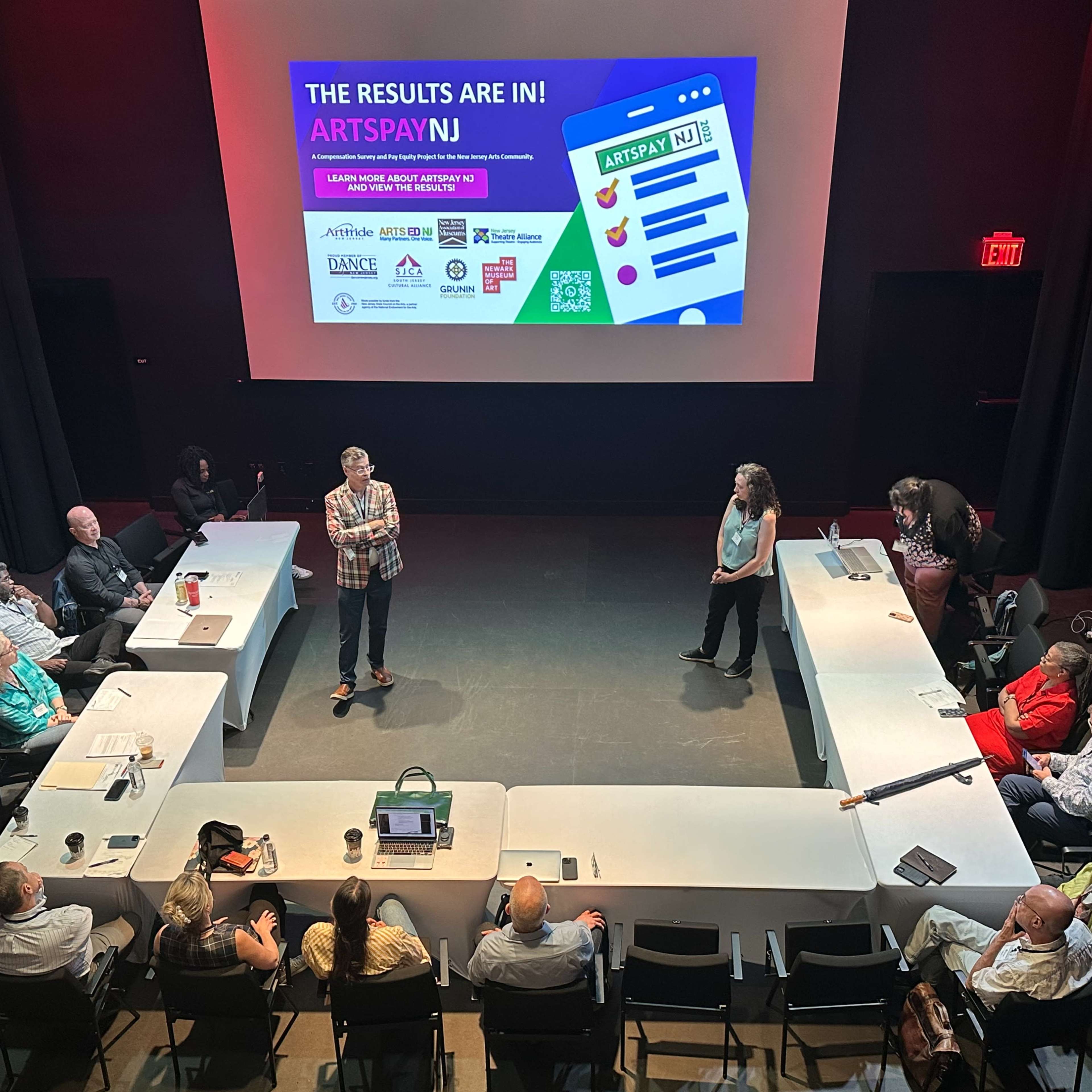 A group of people is seated around a rectangular space in a dimly lit room, while two presenters stand in front of a large screen displaying the text "THE RESULTS ARE IN! ARTSPAY NJ."
