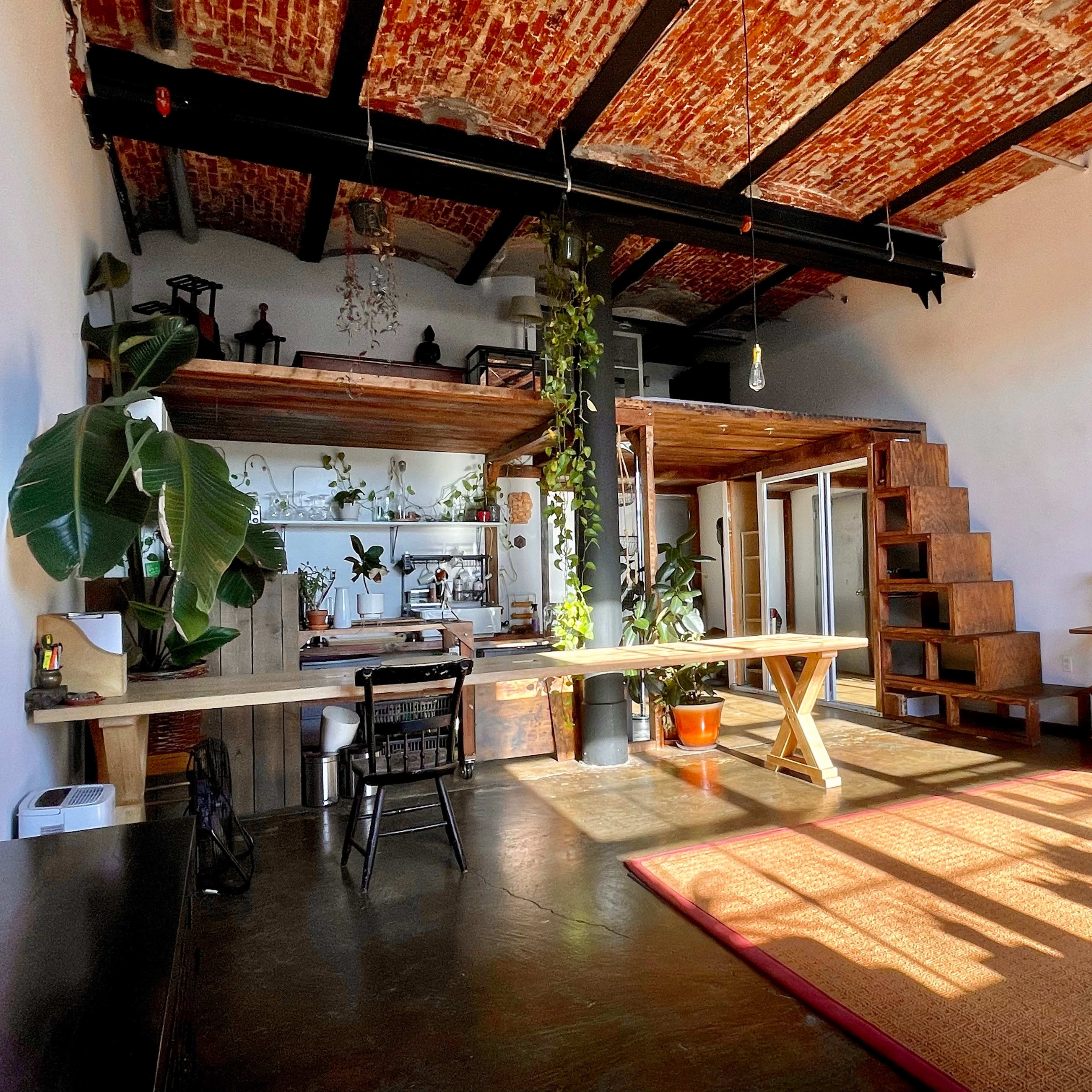 A spacious living area with a wooden loft, plants, and a mix of furniture, including a table, chairs, and a couch.