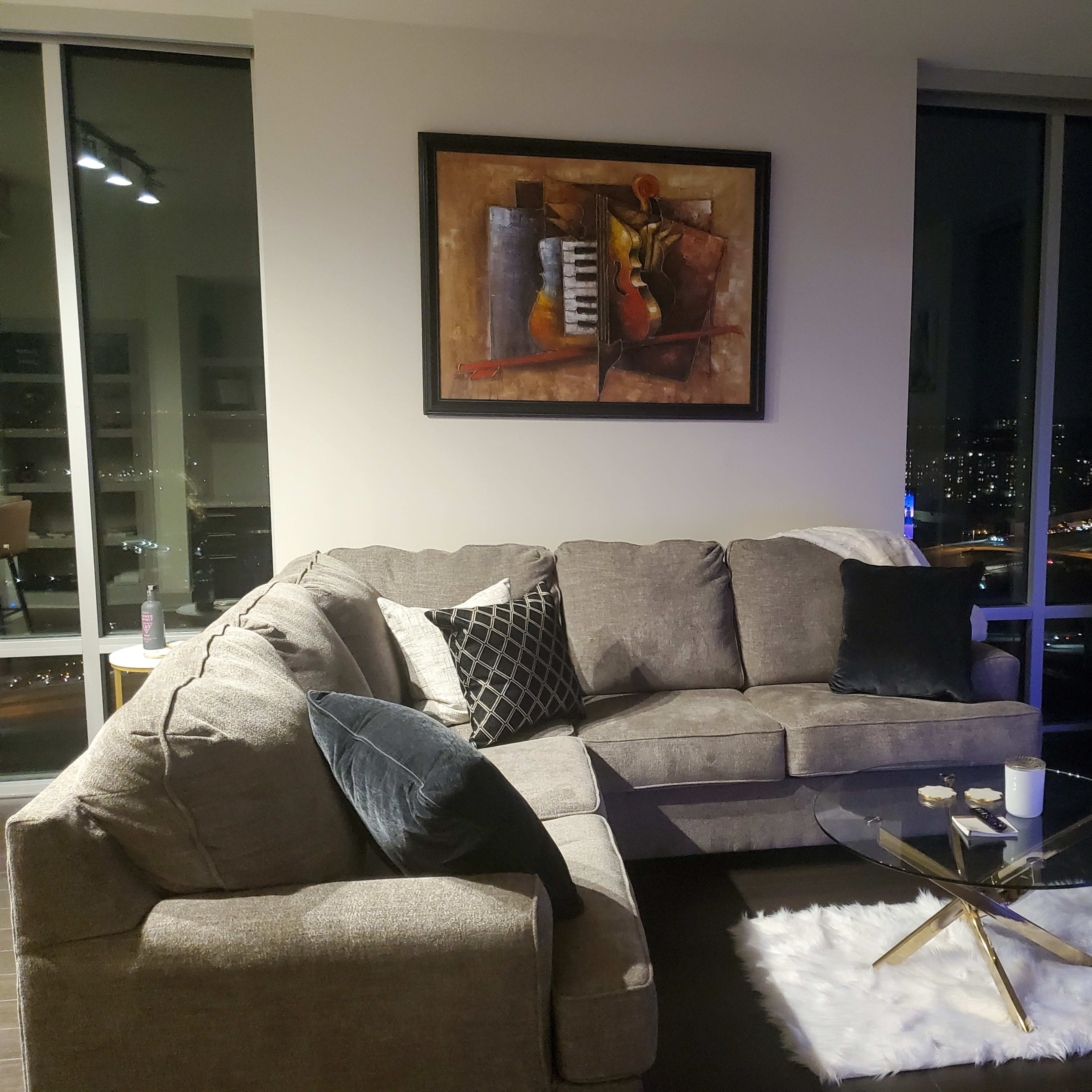 A modern living room features a grey sectional sofa, a glass coffee table on a white rug, and a framed painting on the wall, with city skyline views visible through large windows.