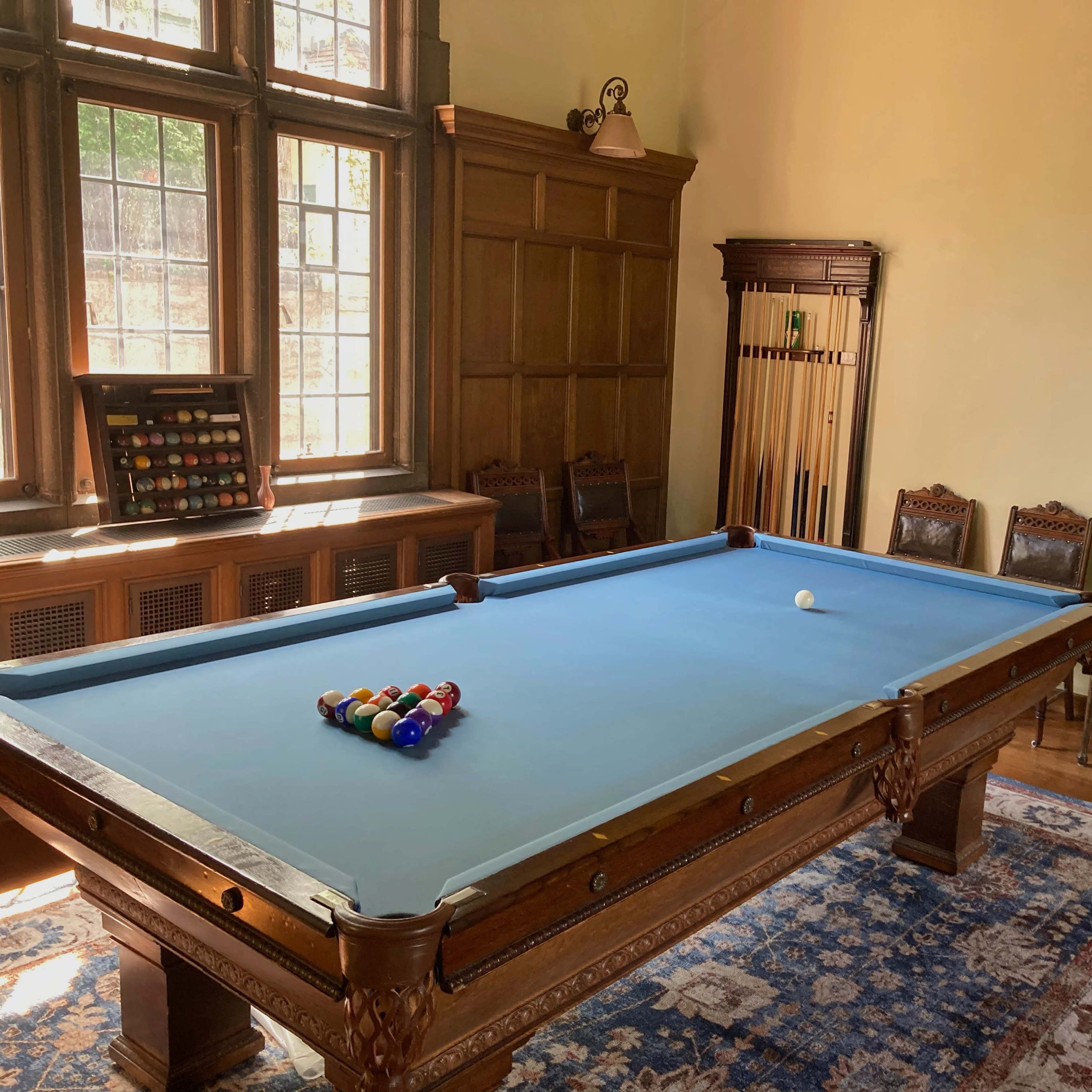 A billiard room featuring a blue felt pool table with a set of colorful balls arranged for a game, surrounded by wooden chairs and a hardwood floor.