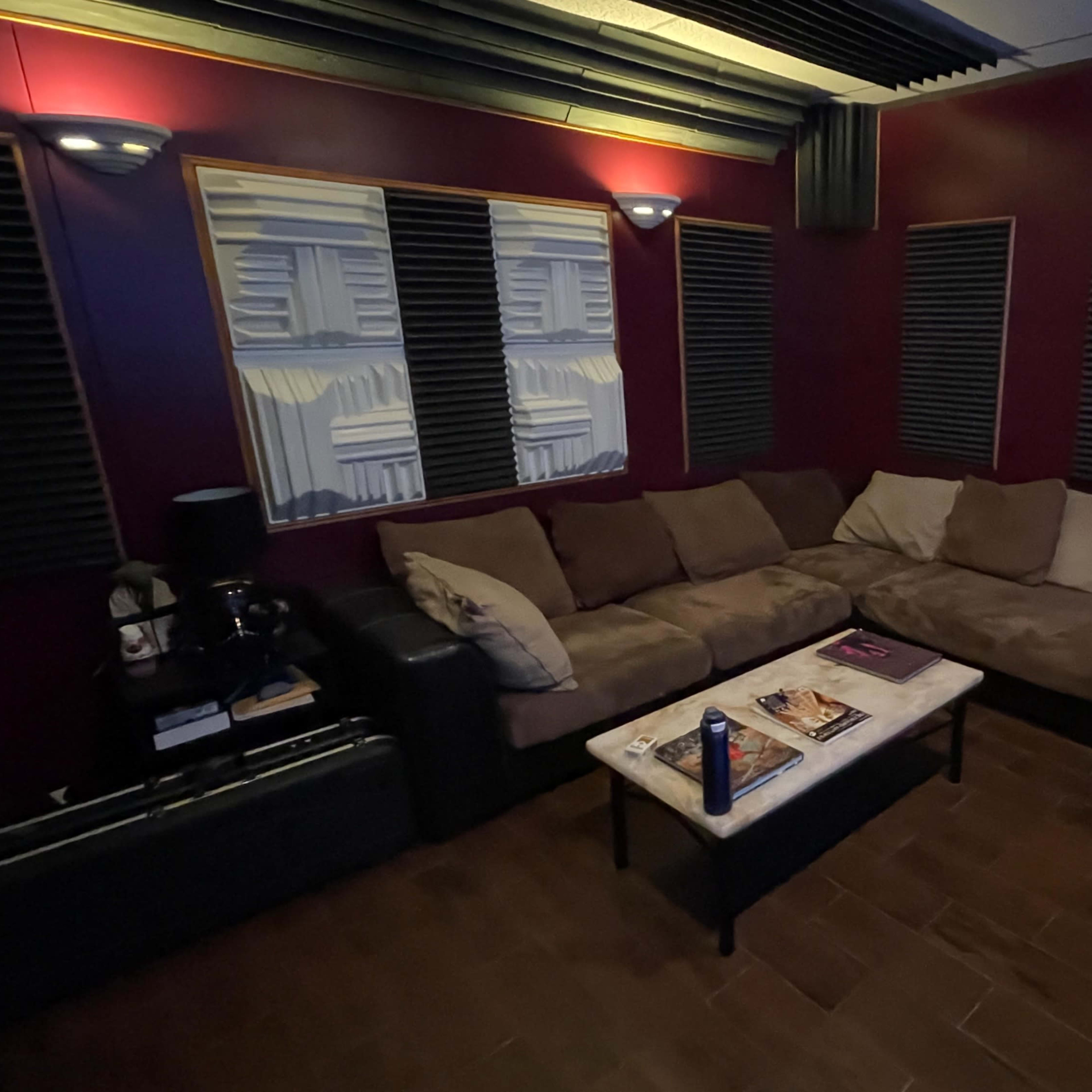 A cozy recording studio with a large brown sectional couch, a coffee table, soundproofing panels on the walls, and a black office chair.
