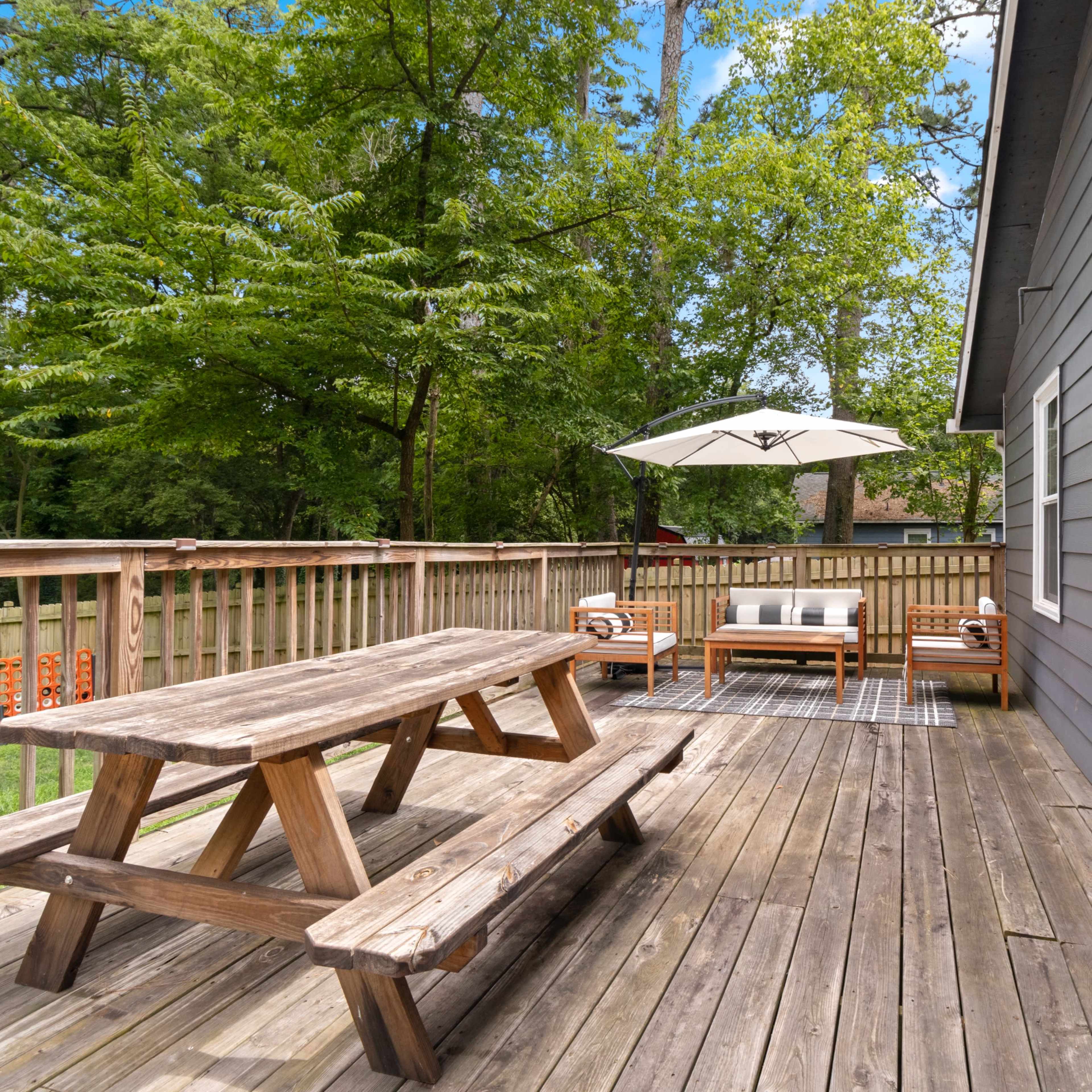 A wooden picnic table and a seating area with a shade umbrella are positioned on a deck surrounded by trees.