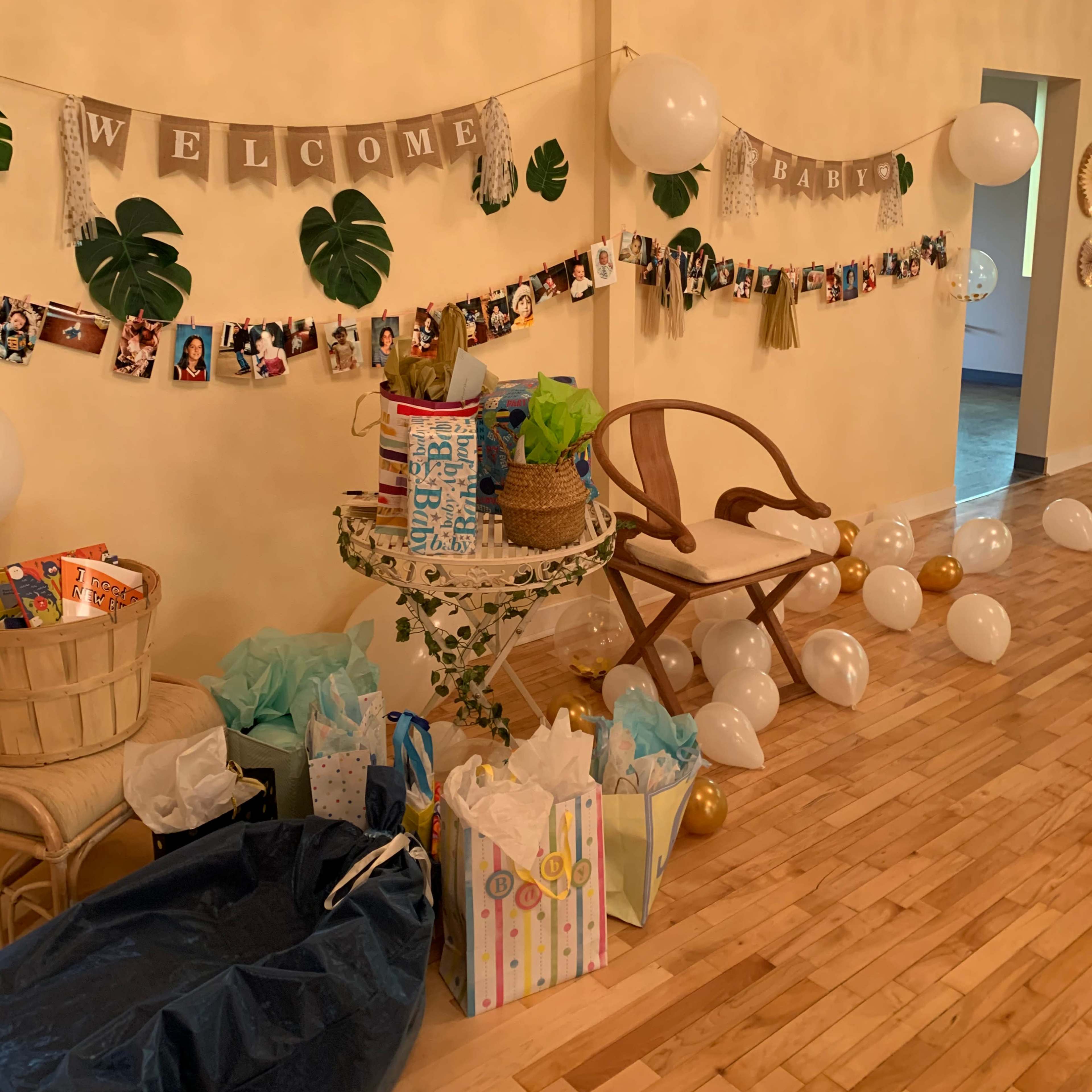A decorated wall with a "Welcome Baby" banner, featuring photos and balloons, is surrounded by a table stacked with gifts and bags.