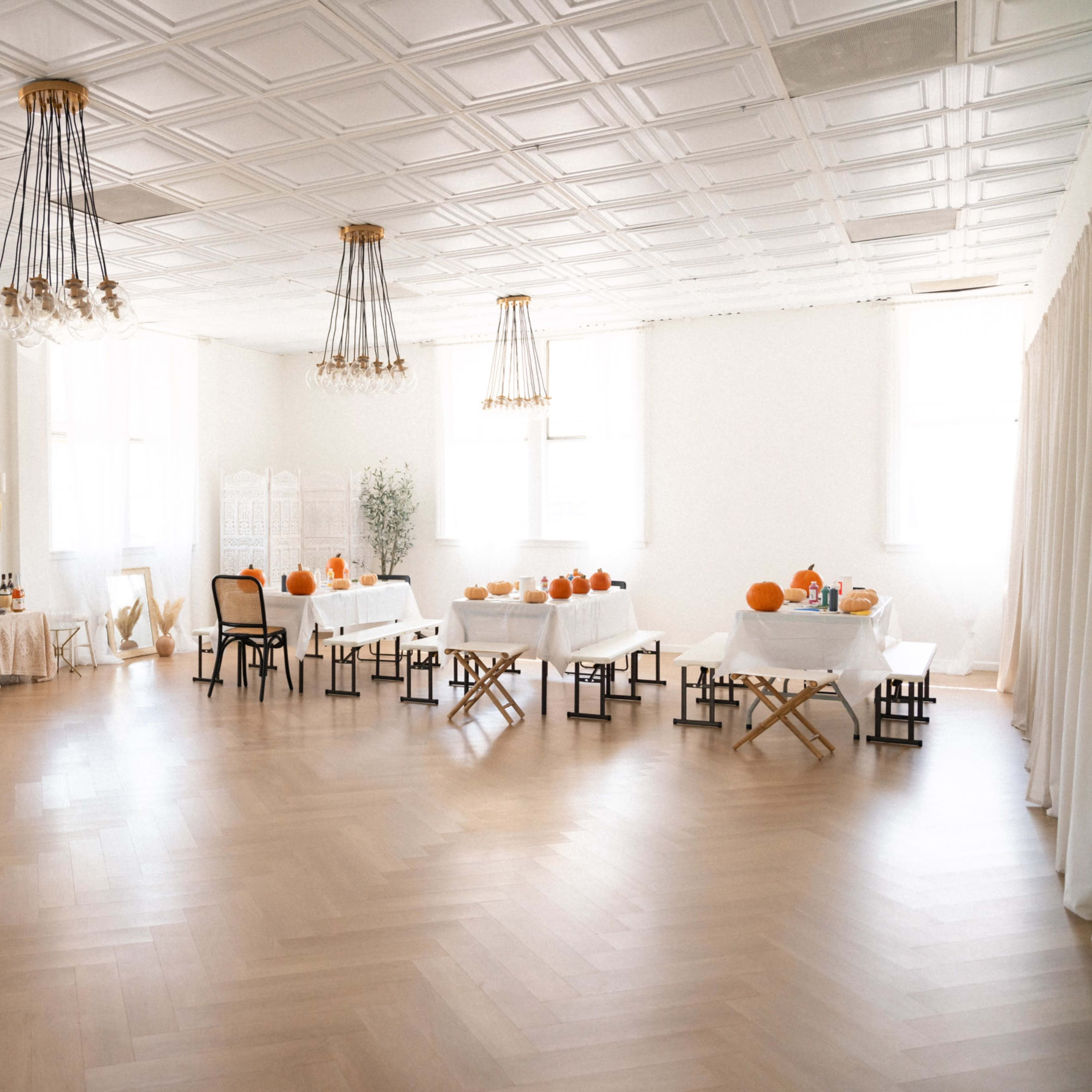 A spacious, well-lit room features several tables adorned with pumpkins and decorative items, surrounded by neutral-colored furnishings and a wooden floor.