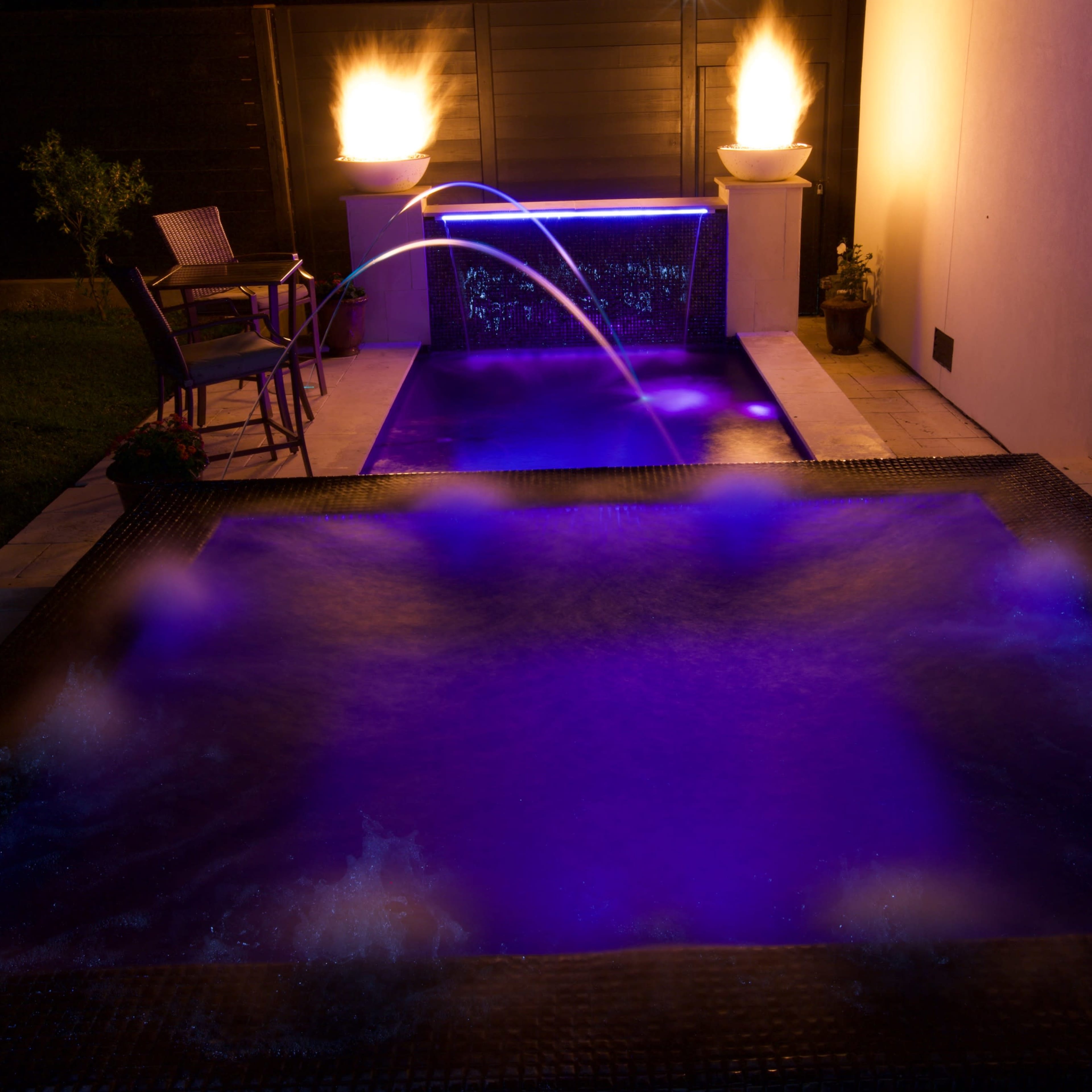 A backyard area with two illuminated water features, one with a fountain and the other with jets, surrounded by a stone deck and a chair.