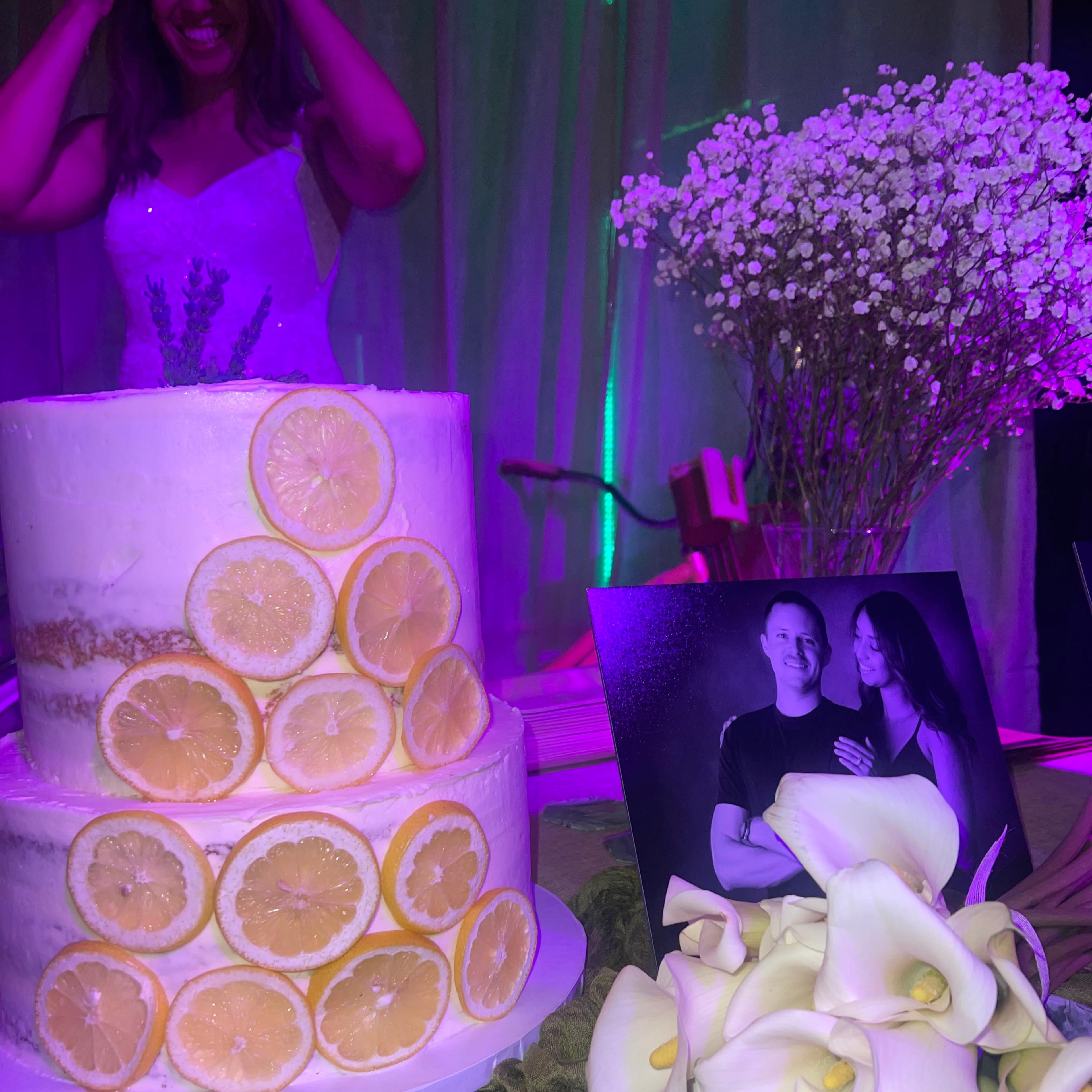 A multi-tiered cake decorated with lemon slices stands on a table next to a black and white photo of a couple, with flowers and greenery in the background.