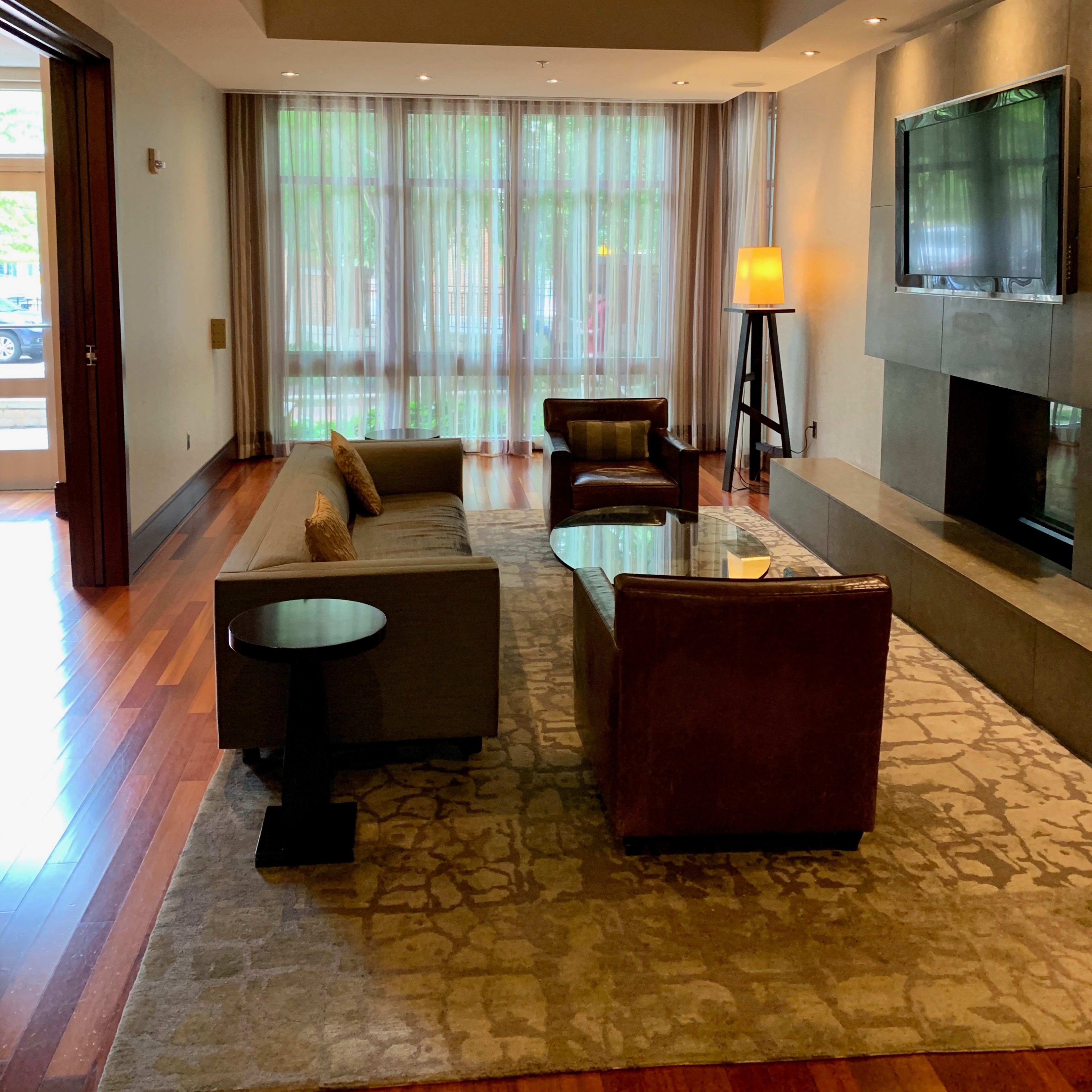 A modern living room with a sofa, a chair, and a glass coffee table, illuminated by soft light from a floor lamp and large windows covered with sheer curtains.
