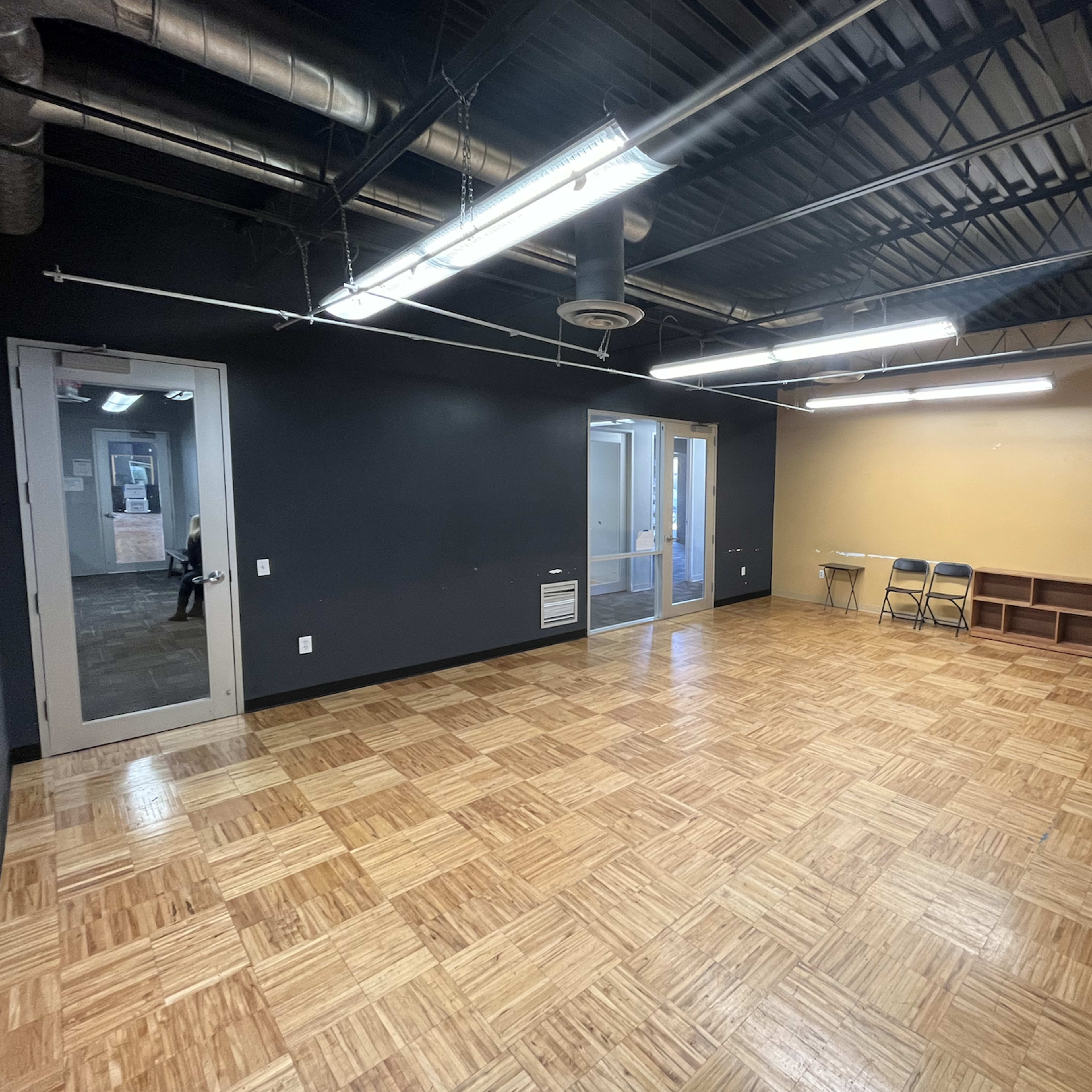 A spacious, empty room with a wooden floor, black walls, and a large windowed door leading to an adjacent area.