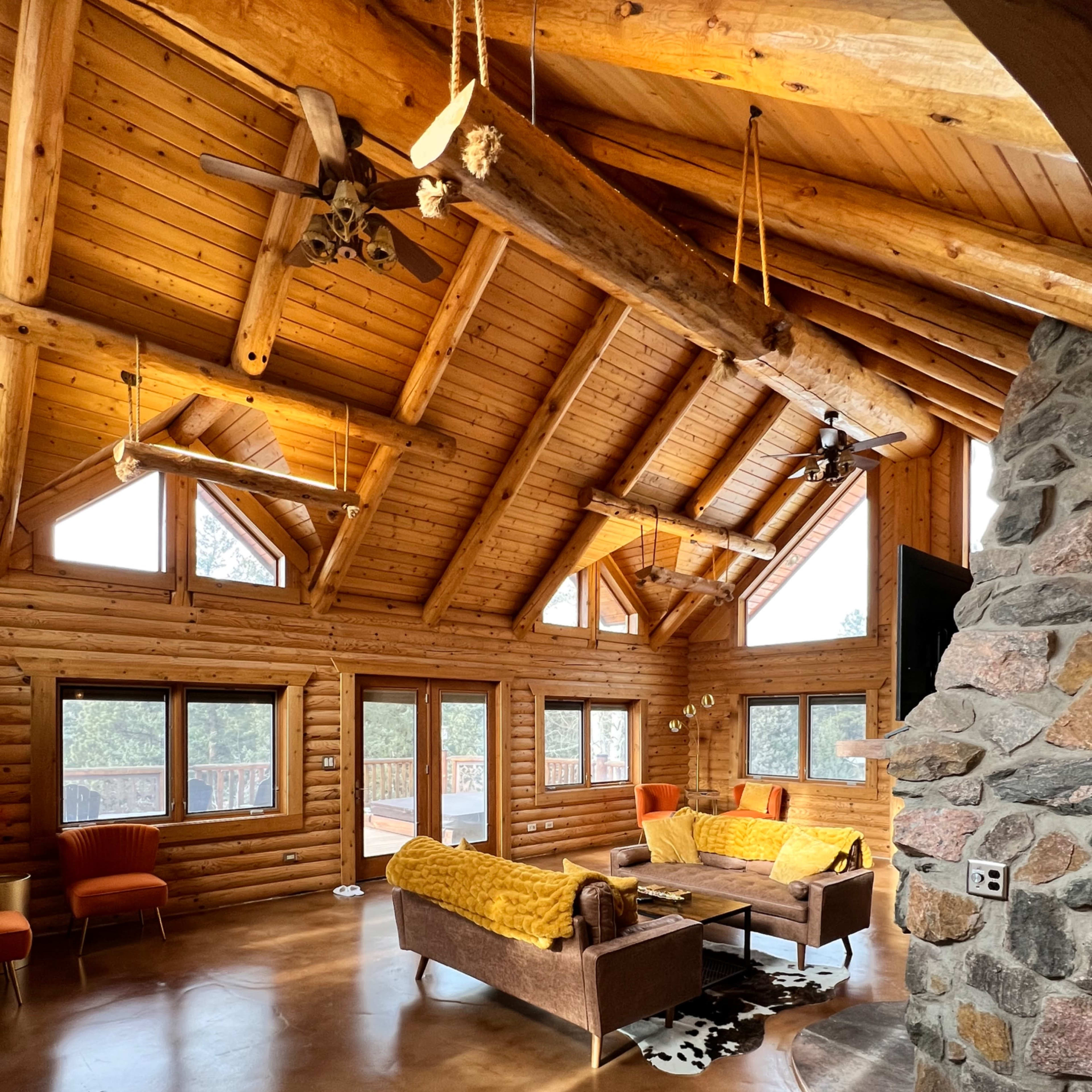 The interior of a log cabin features a high, wooden-beamed ceiling, large windows, and rustic furniture arranged around a stone fireplace.