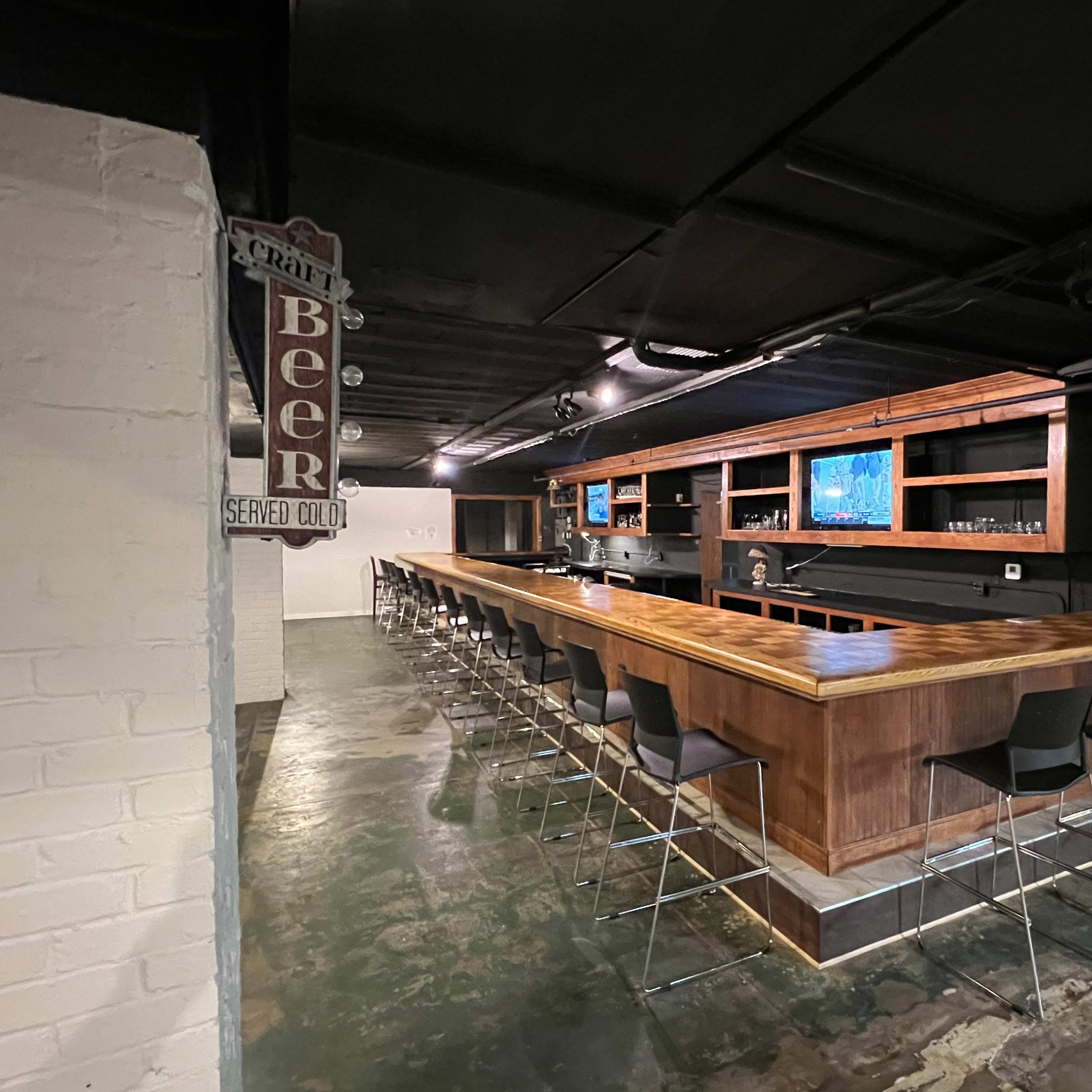 A modern bar with a long wooden counter and high black stools, set in an industrial-style space with exposed walls and dim lighting.