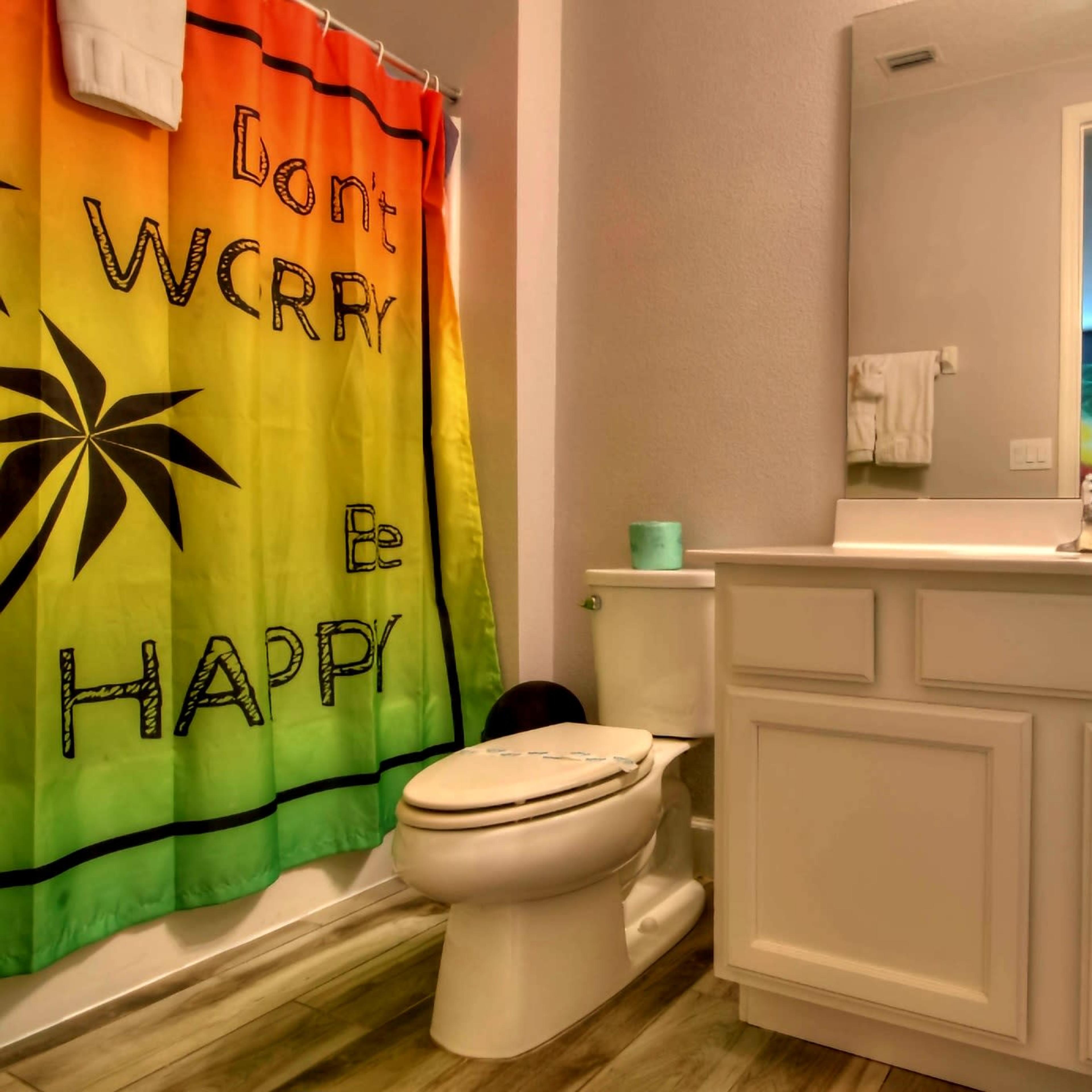 A bathroom features a colorful shower curtain with the text "Don't Worry Be Happy," a white toilet, and a vanity with a mirror.