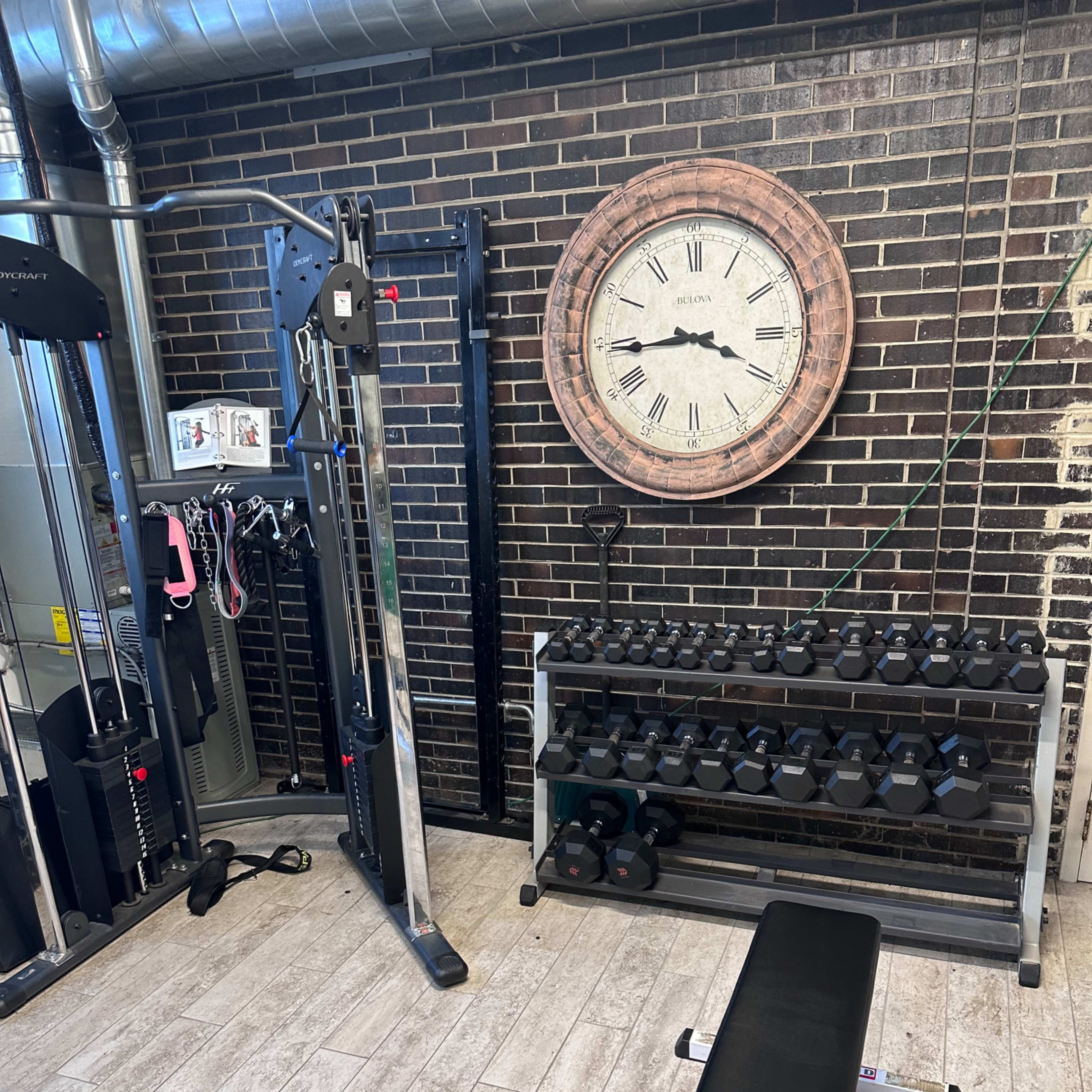 A home gym area with a weight bench, dumbbell rack, and a cable machine against a brick wall, complemented by a large wall clock.