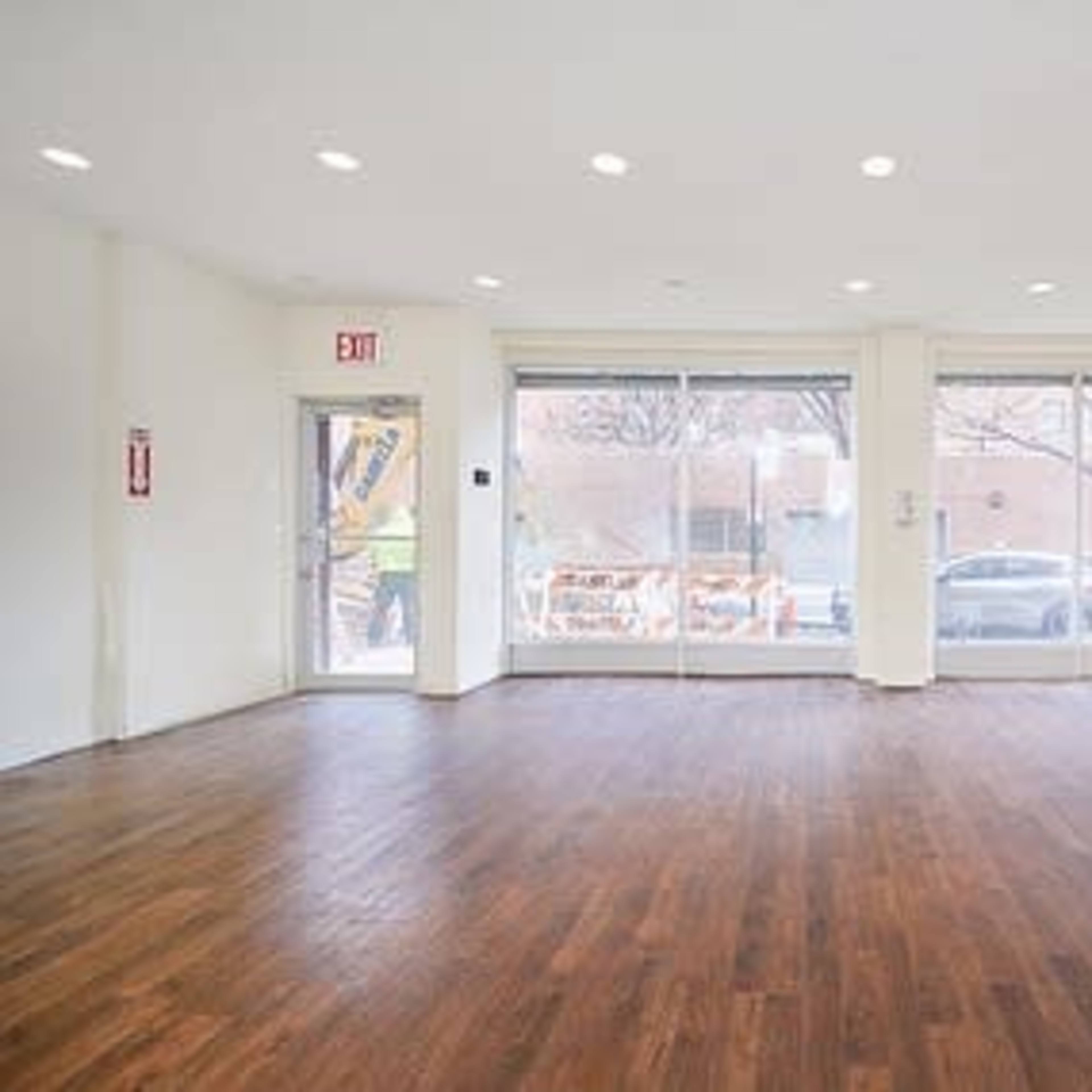 An empty, well-lit room with wooden flooring and large windows facing a street.
