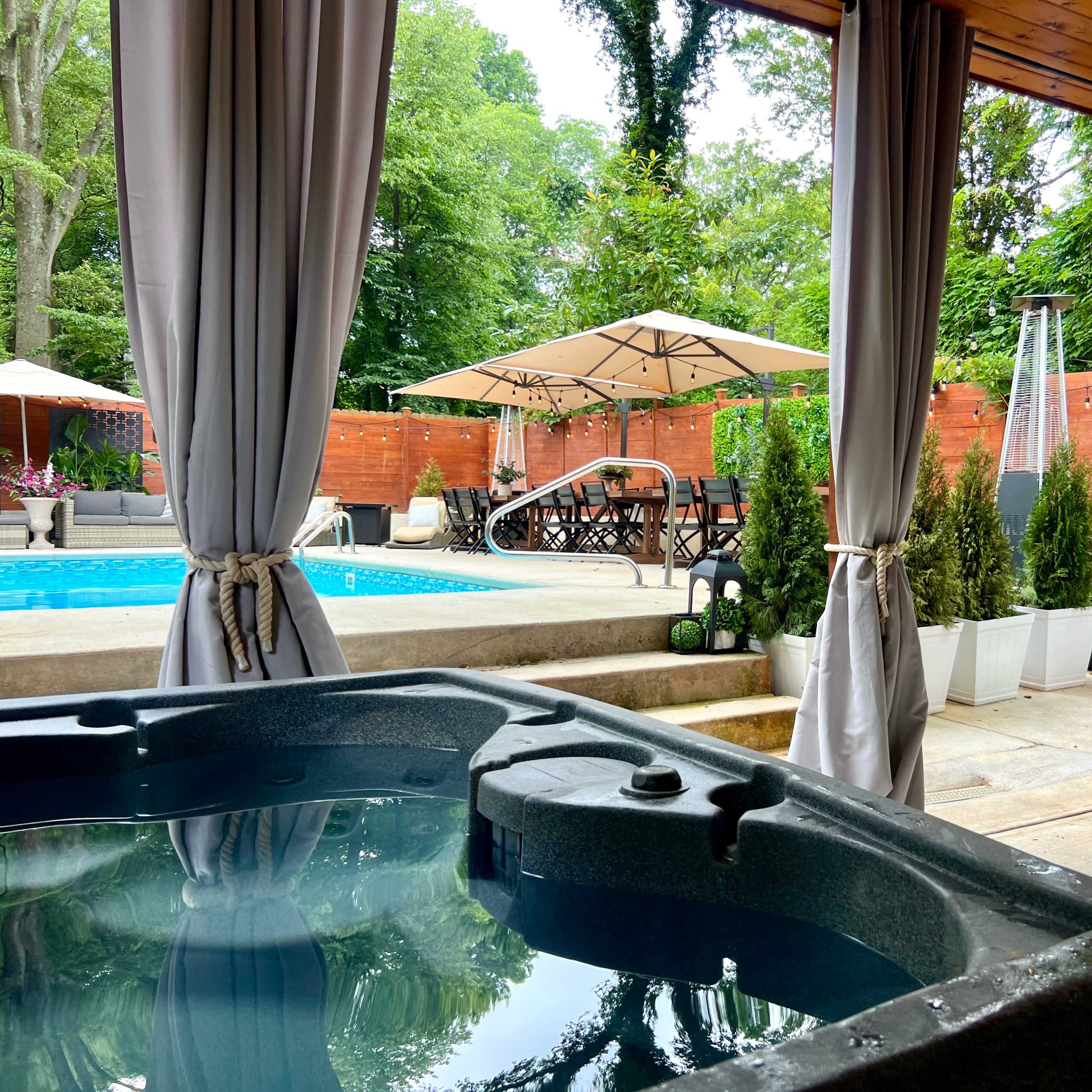 A backyard scene with a hot tub in the foreground, a swimming pool in the background, and various outdoor furniture and greenery.