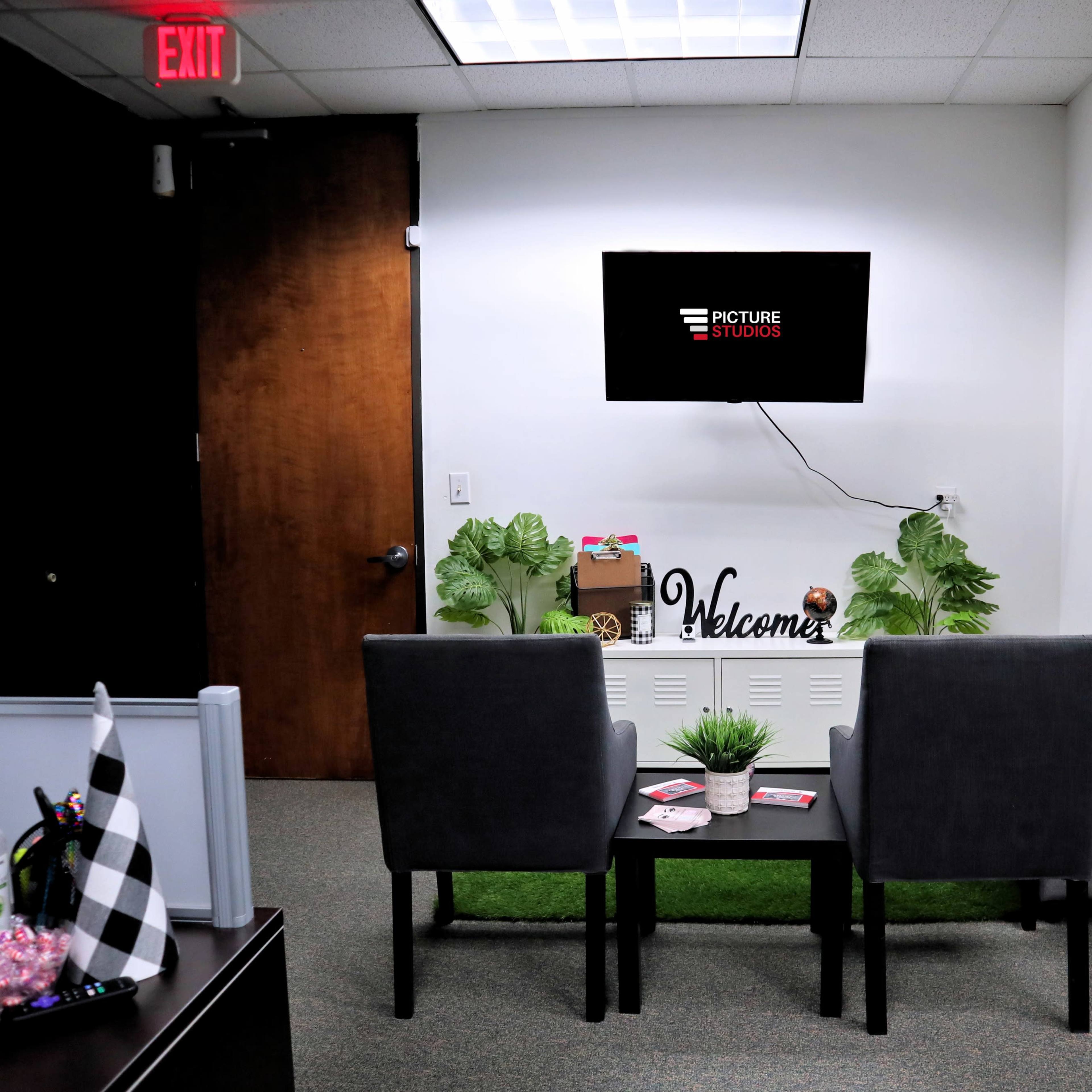 A small waiting area with two chairs, a table, and a television mounted on the wall, featuring decorative plants and a welcome sign.