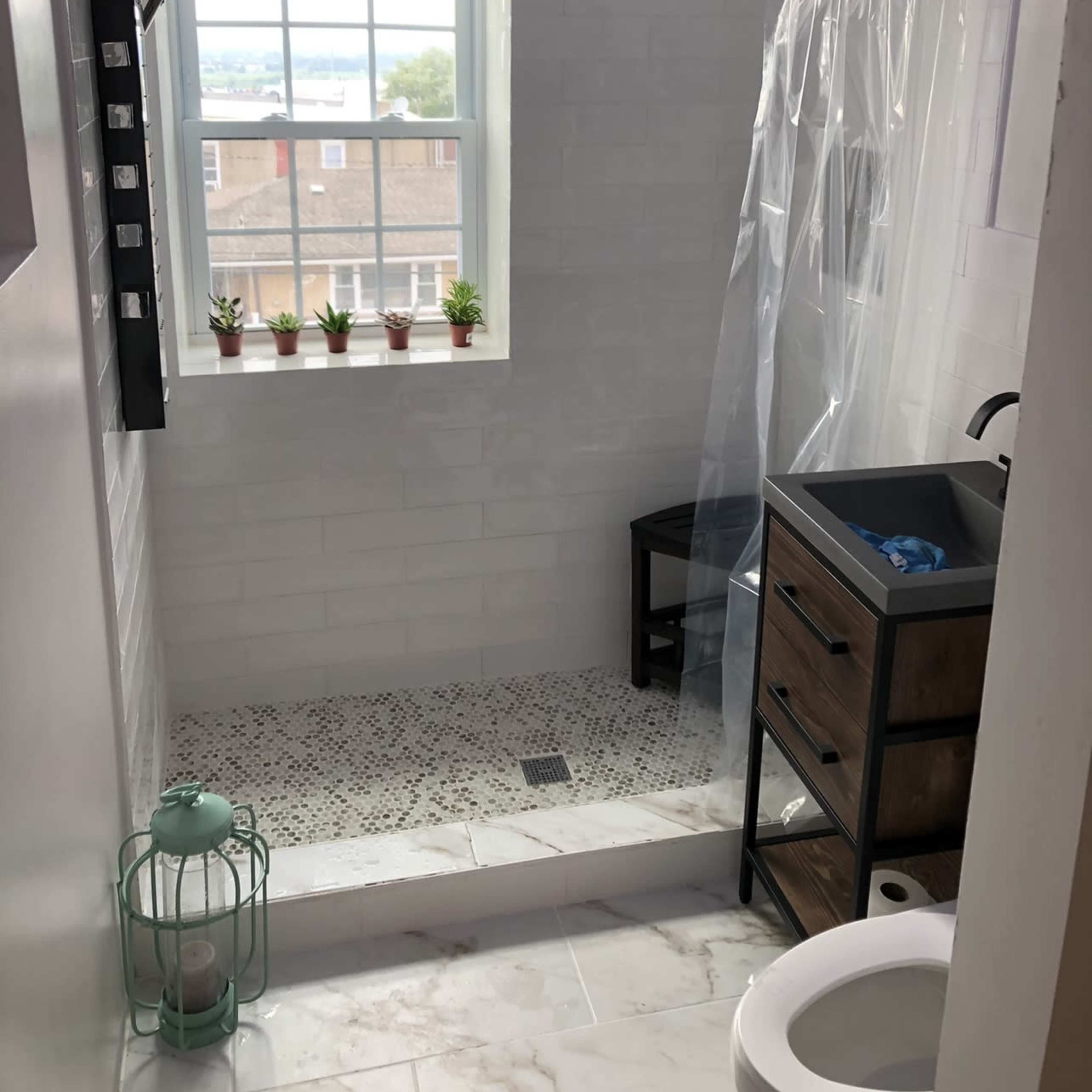 A modern bathroom with a walk-in shower featuring a clear curtain, a small sink cabinet, and a window with potted plants.
