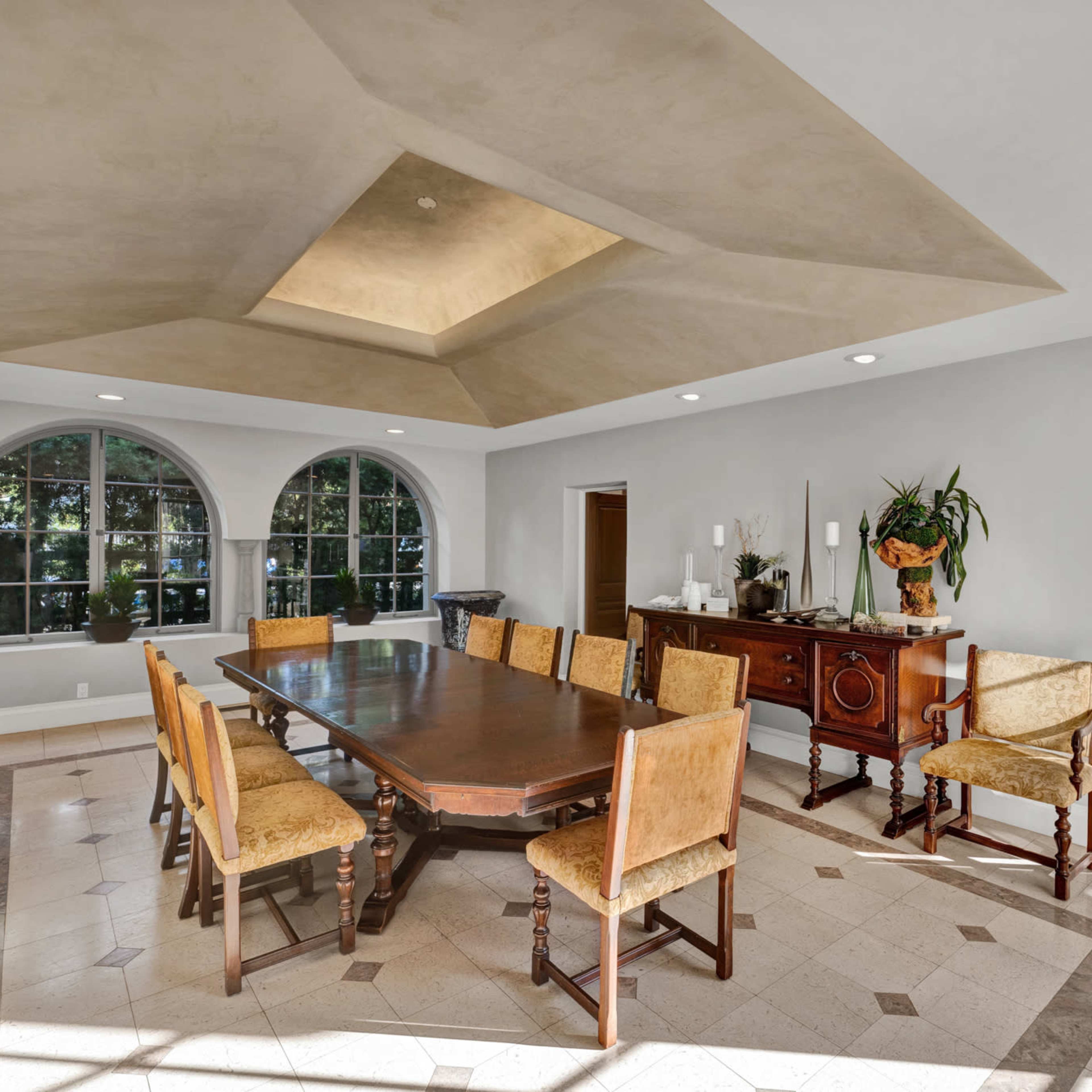 A spacious dining room features a large wooden table surrounded by upholstered chairs and windows that overlook a garden.
