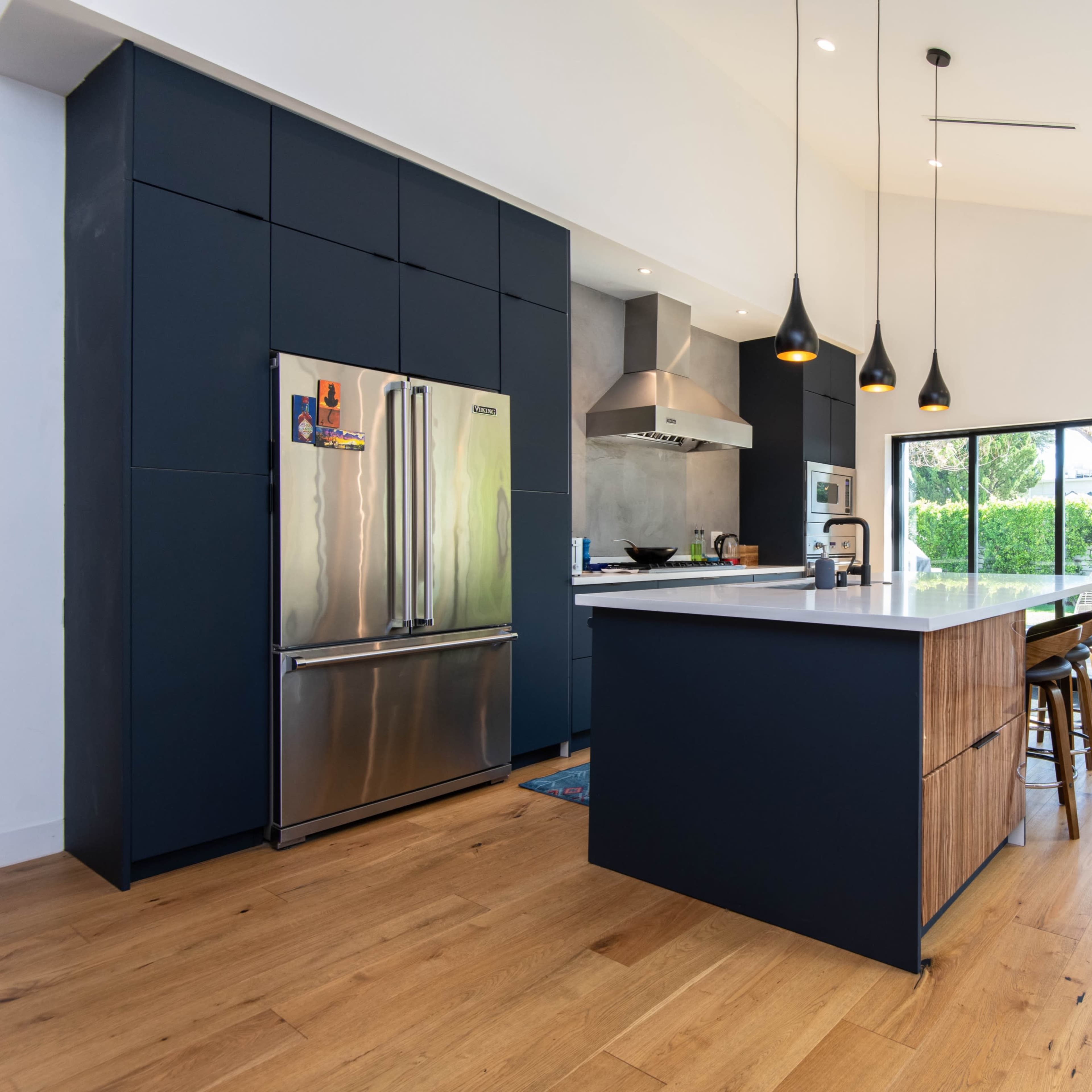 A modern kitchen with dark cabinets, a stainless steel refrigerator, and an island with wooden accents, leading to a patio area through large sliding glass doors.