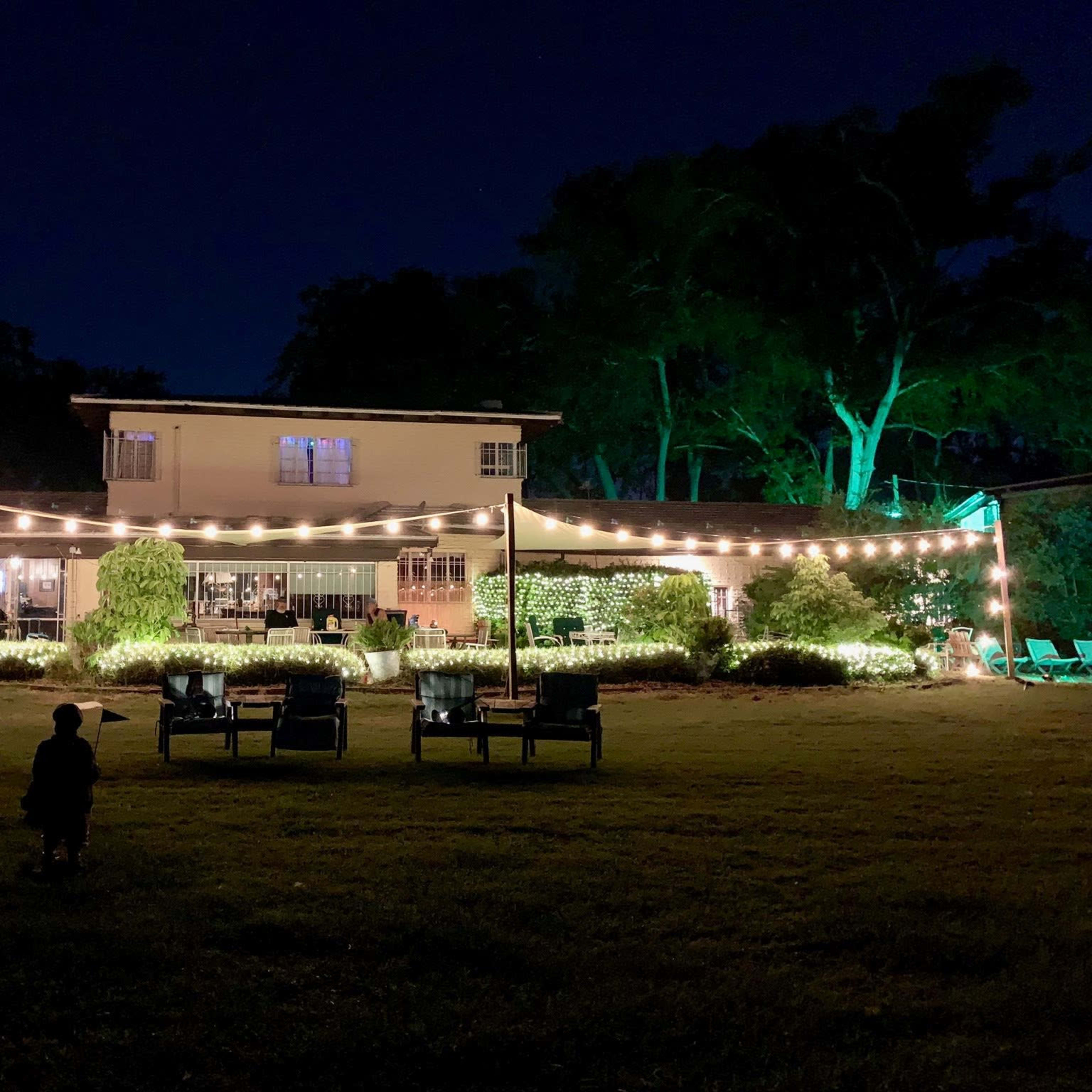 A house is illuminated by string lights in a yard at night, with chairs arranged in front and trees in the background.
