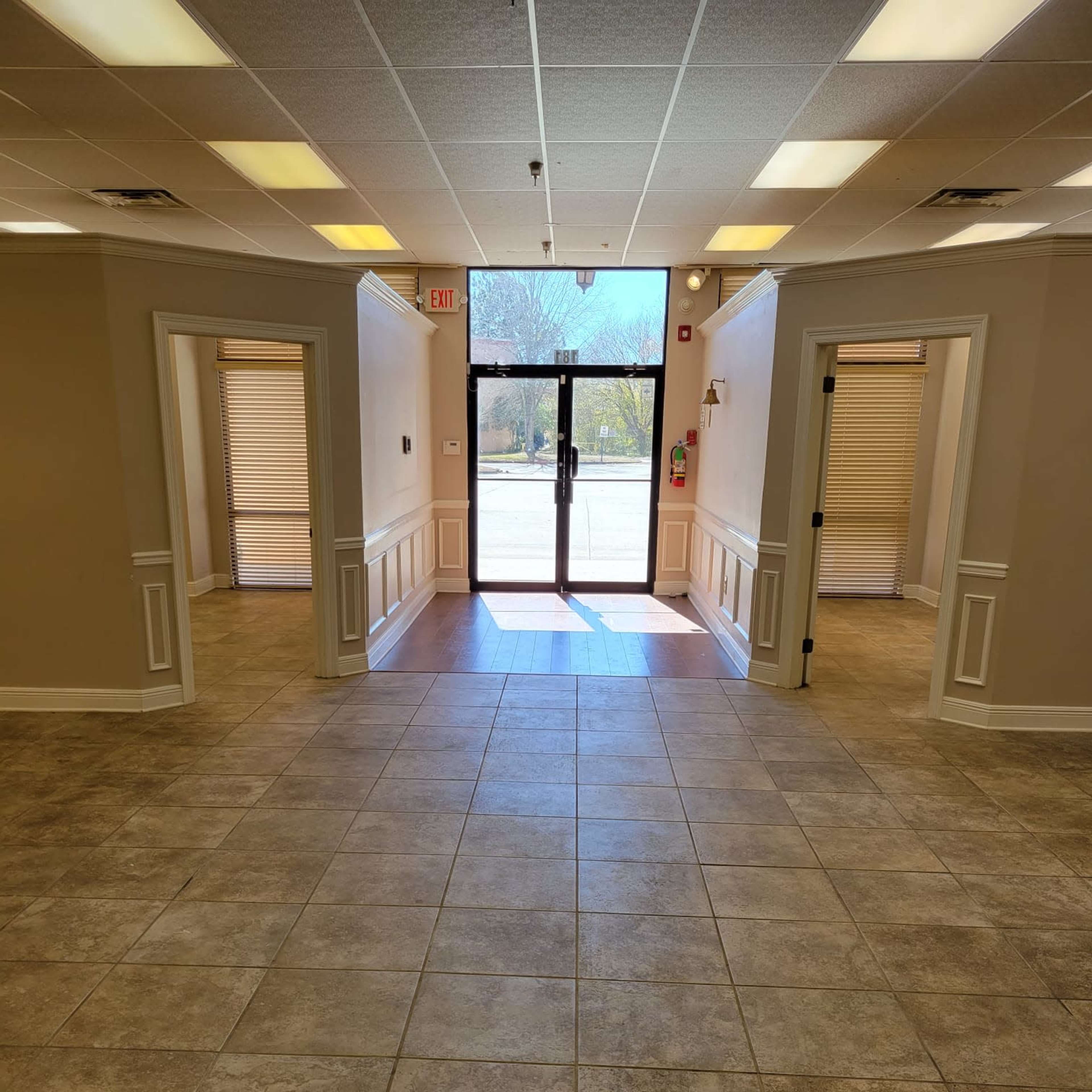 A spacious corridor with tile flooring leading to a glass entryway, flanked by doorways on either side.