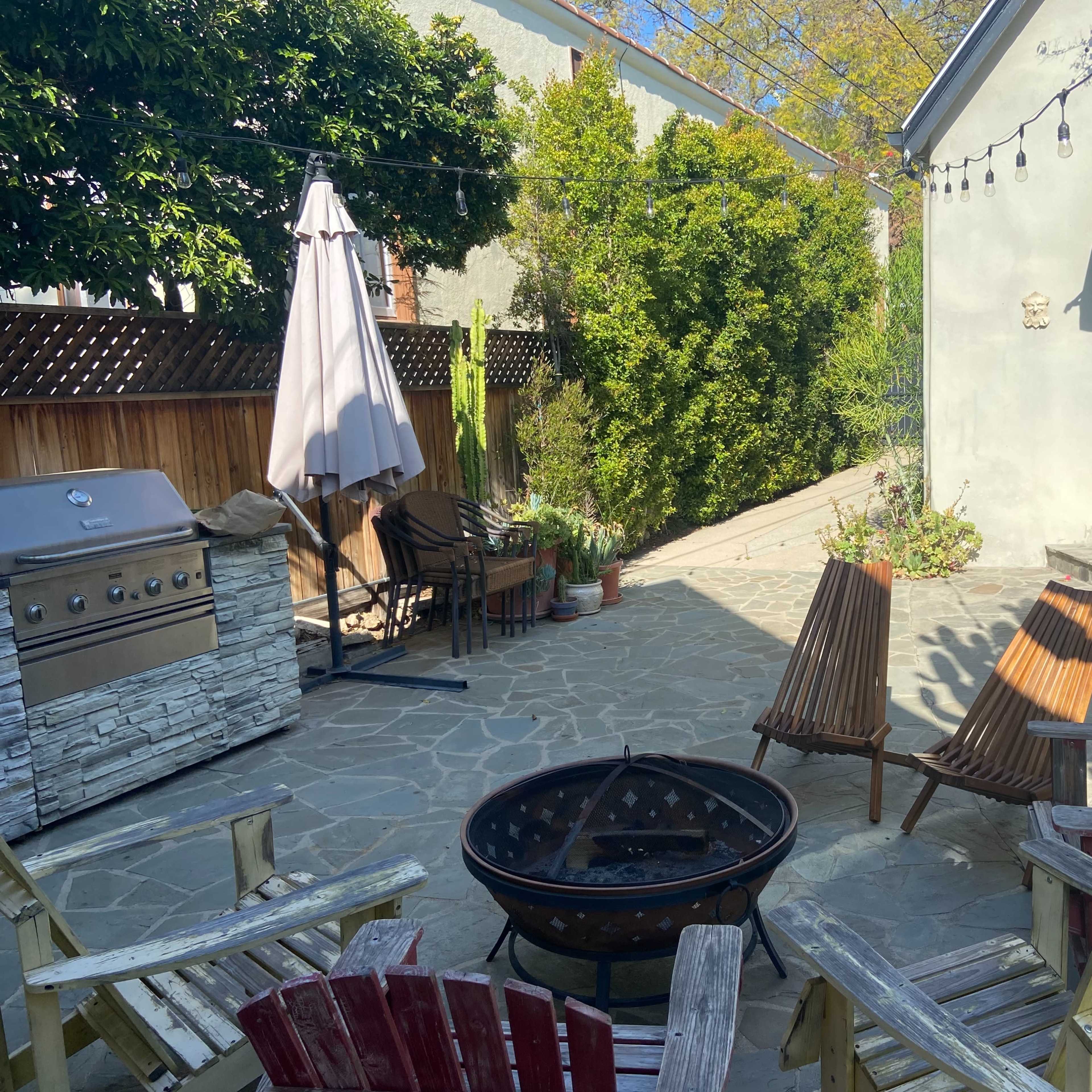 A stone patio with a fire pit, several wooden chairs arranged around it, and a stainless steel grill nearby, framed by greenery and a light-colored house.
