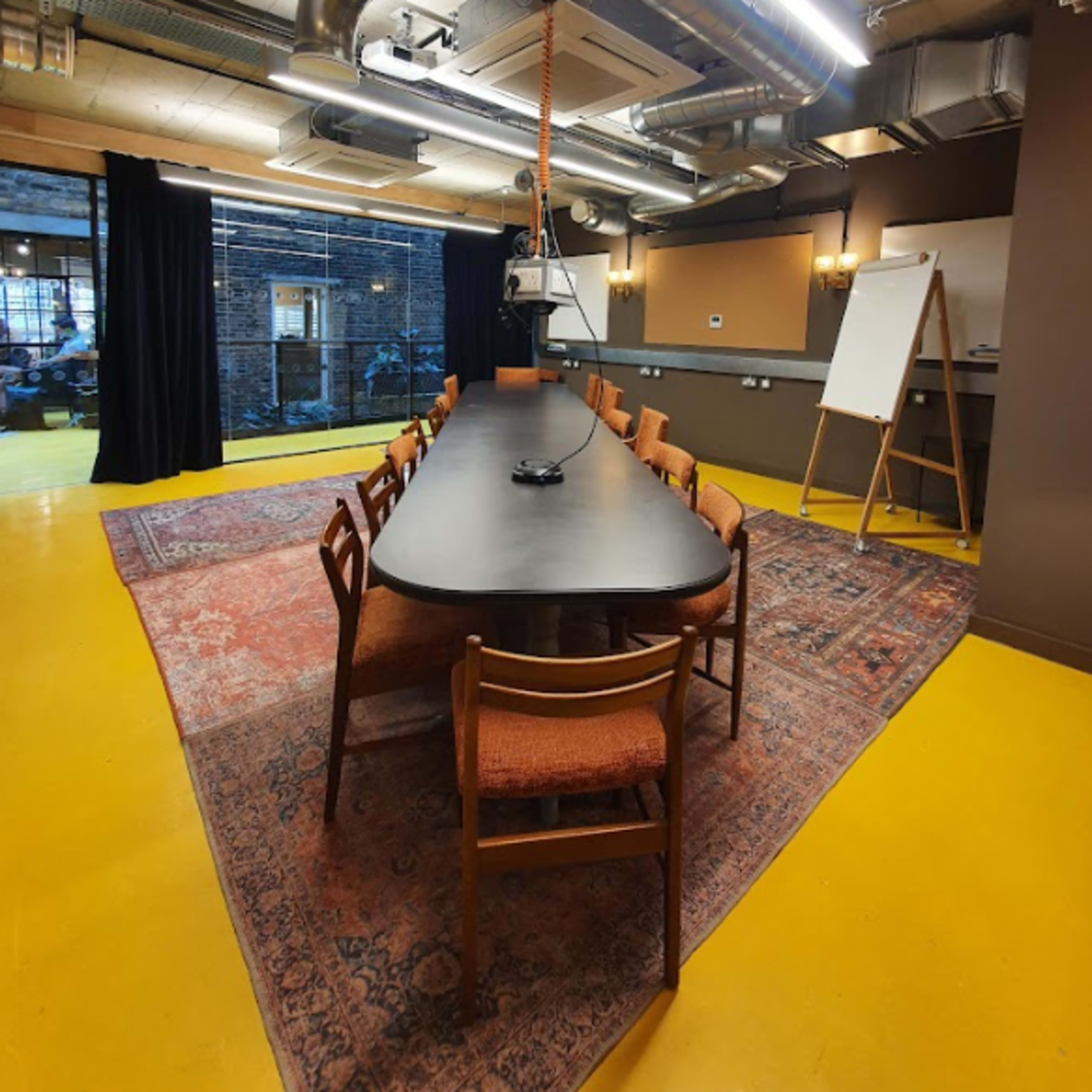 A long rectangular table with wooden chairs is set in a meeting room featuring industrial-style decor and a yellow floor.