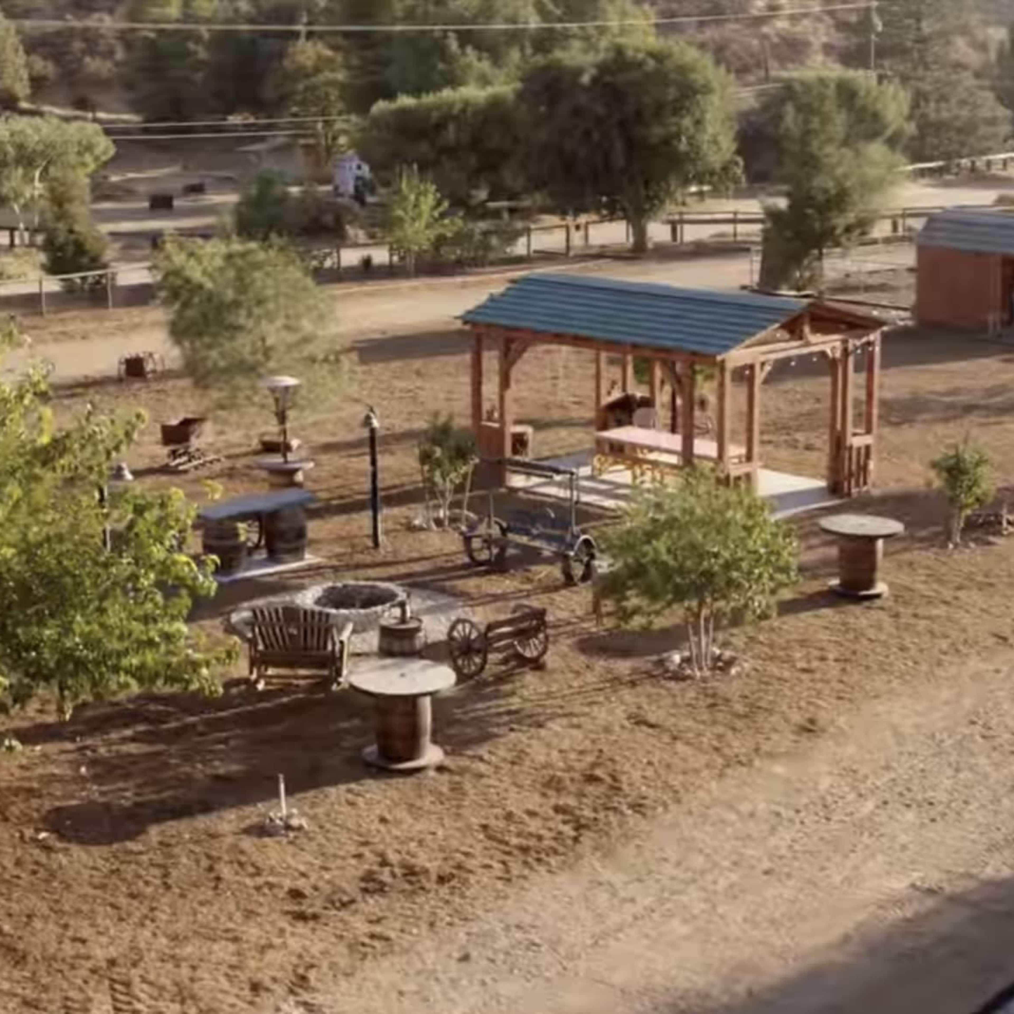 An outdoor area with wooden structures, including a covered gazebo, surrounded by trees and garden furniture.