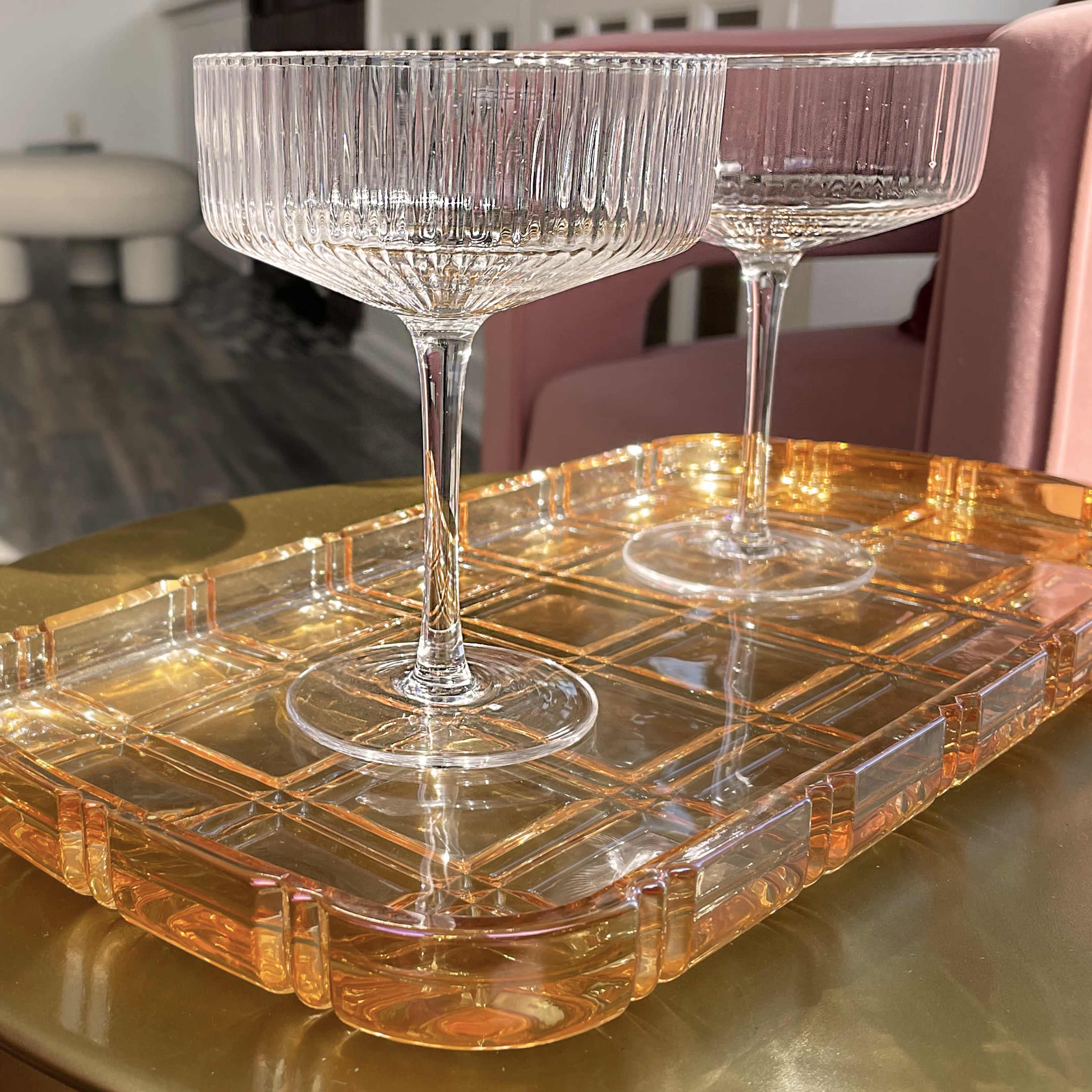 Two crystal champagne coupes sit on a translucent orange tray atop a gold table.