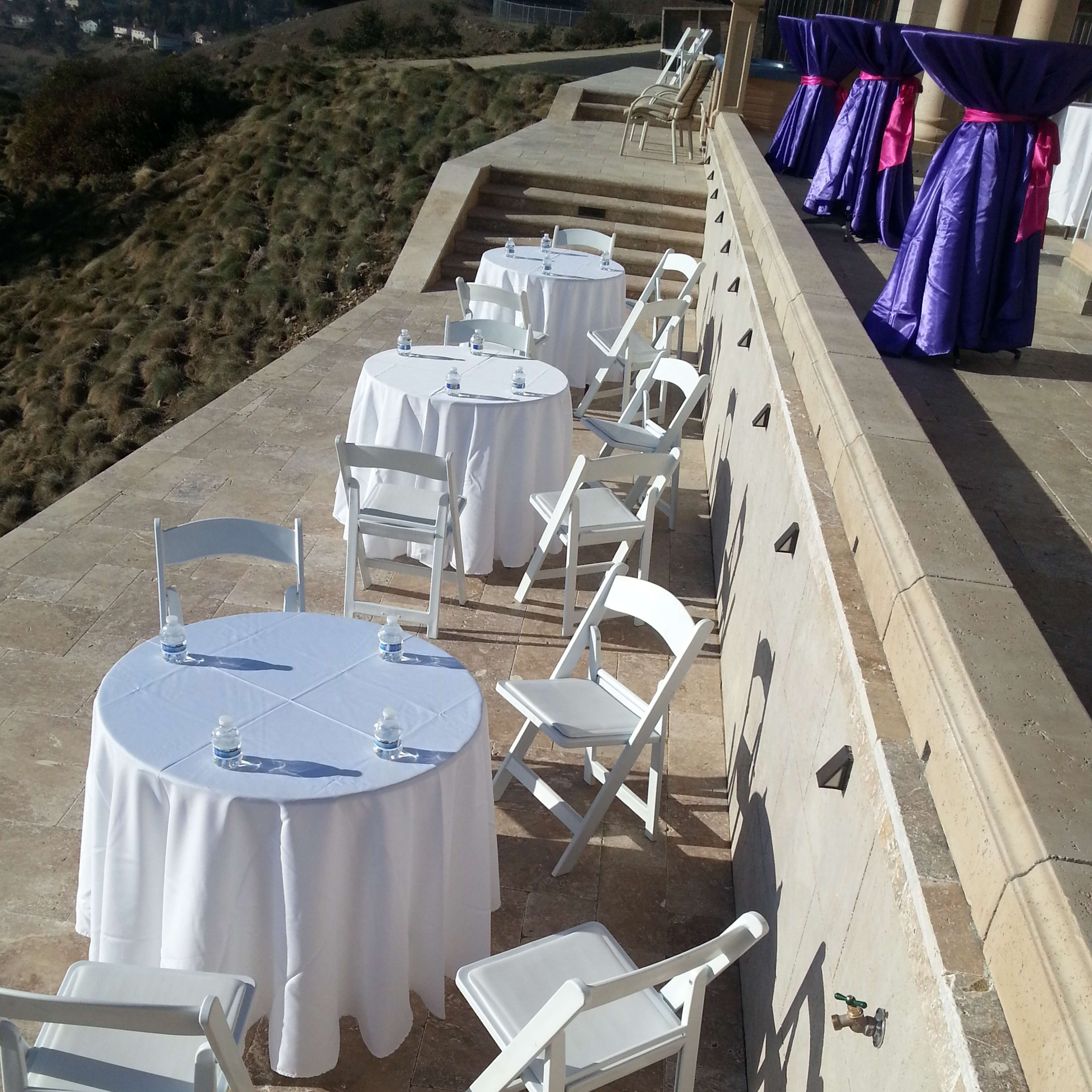Several round tables with white tablecloths and plastic chairs arranged on a stone patio overlooking a hillside.